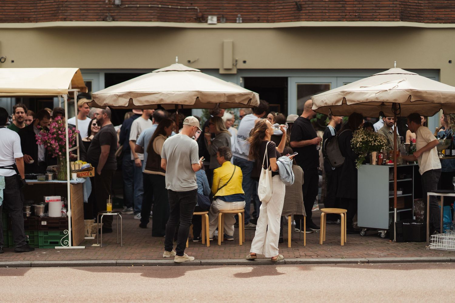 Crowd of people socializing at an outdoor market event with large patio umbrellas and vendor stalls.