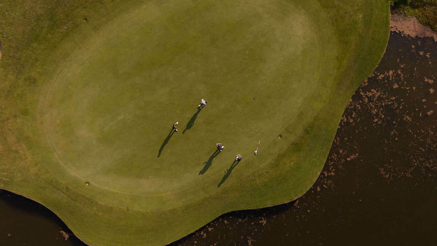 Aerial view of three golfers casting long shadows on a lush green golf course beside a water hazard.