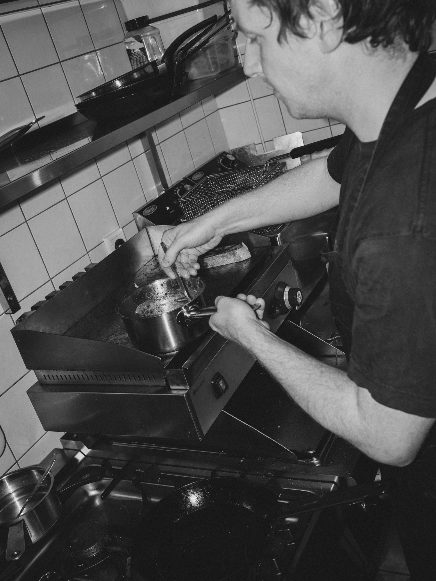 Black and white photo of a chef cooking on a professional stovetop in a tiled kitchen.