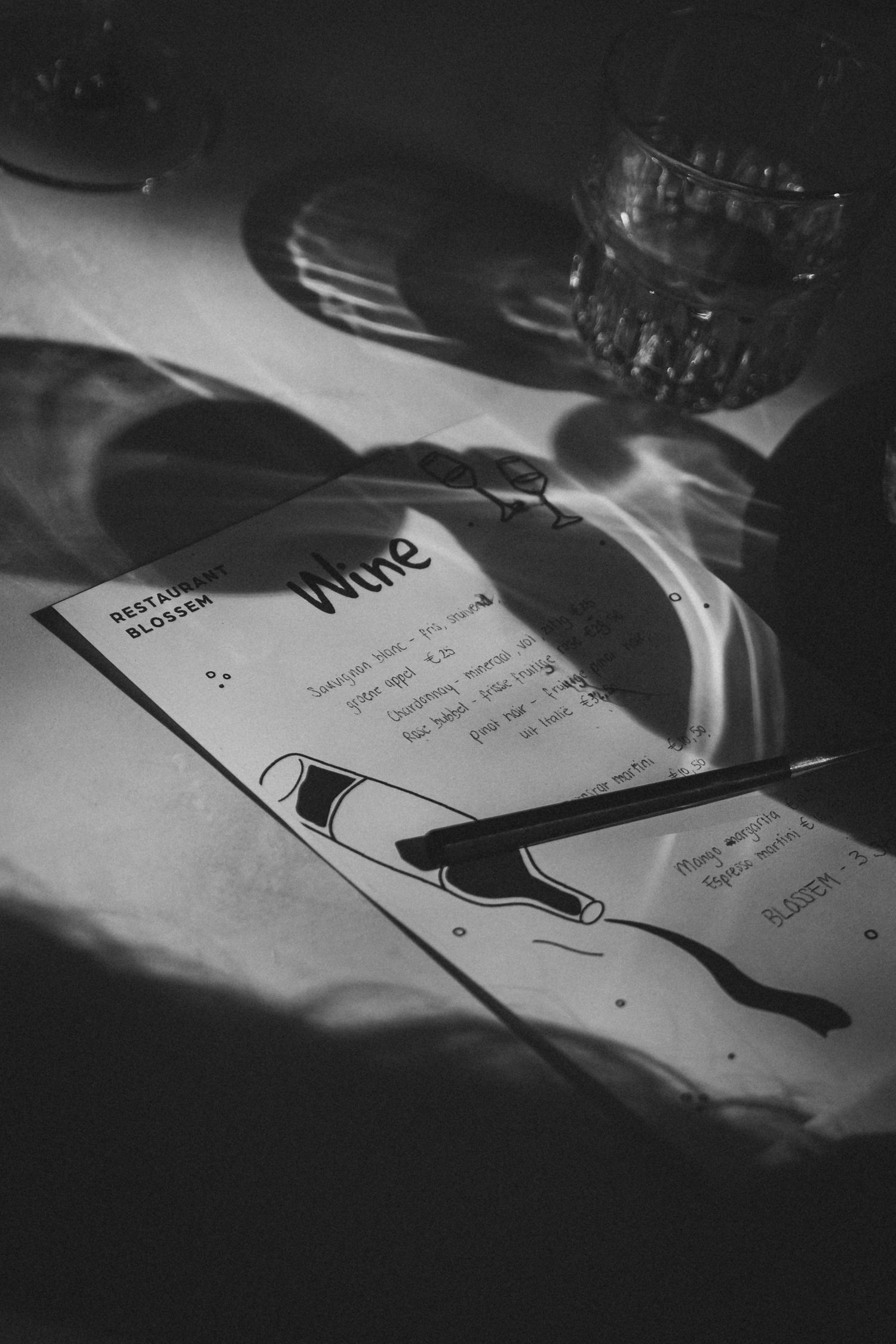 Black and white photo of a wine menu with a pen on a restaurant table with glassware.