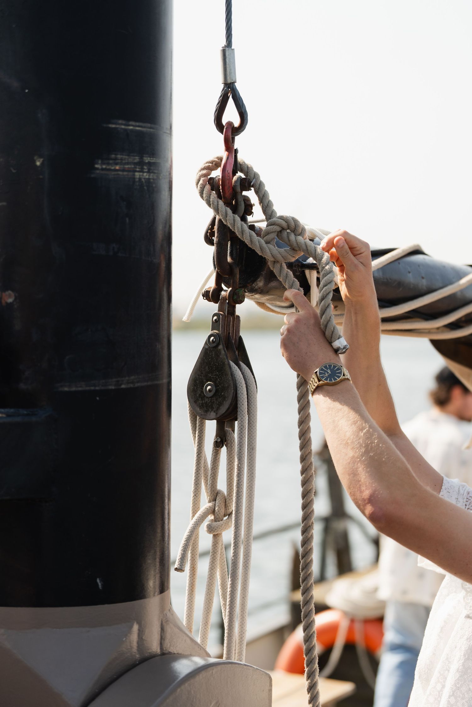 Sailor's hands adjusting rigging ropes on a wooden sailing boat mast with pulley blocks on a sunny day.