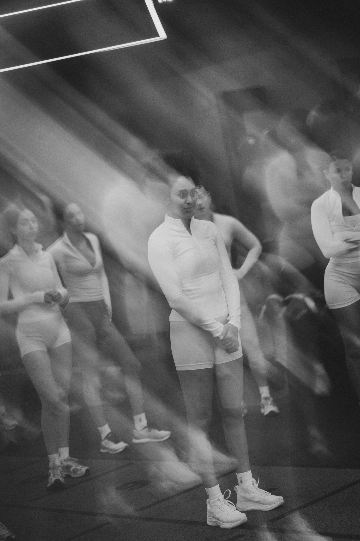 Black and white motion blur photo of athletes in white sportswear exercising in a gym under bright lights.