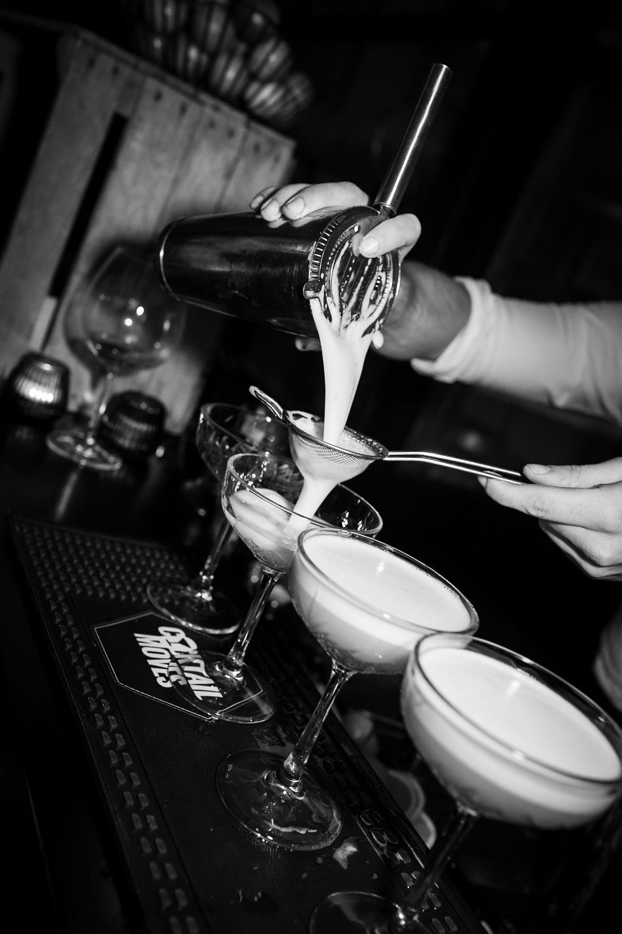 Bartender pouring creamy cocktails into martini glasses at a bar in black and white photography.