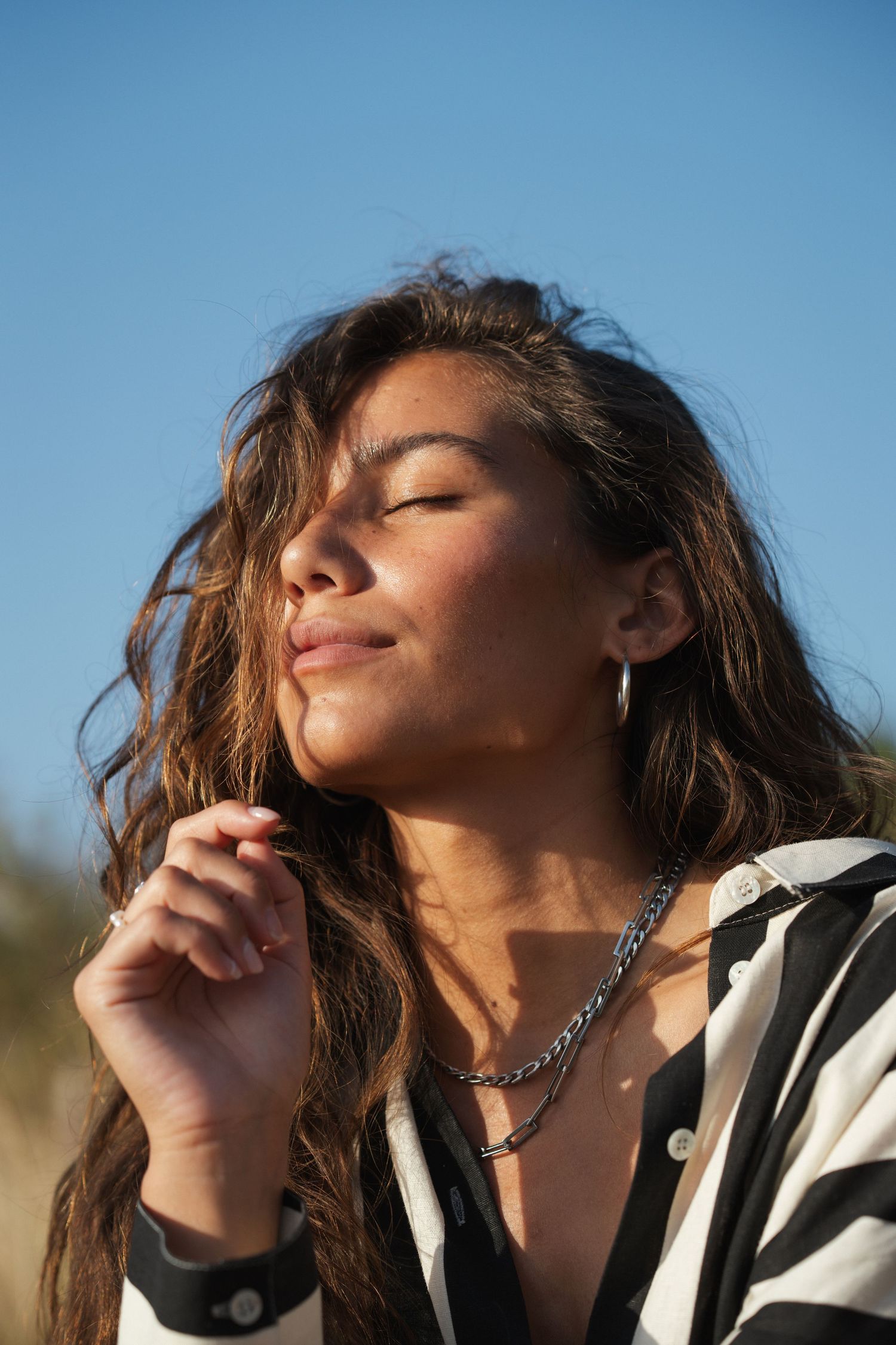 Young woman with wavy hair, eyes closed, enjoying sunlight outdoors, wearing striped shirt and silver chain necklace.