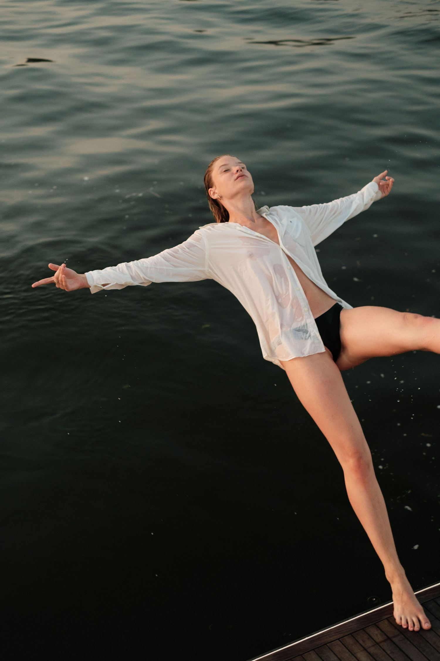 Woman in wet white shirt and black shorts posing on yacht deck with arms outstretched over calm water.