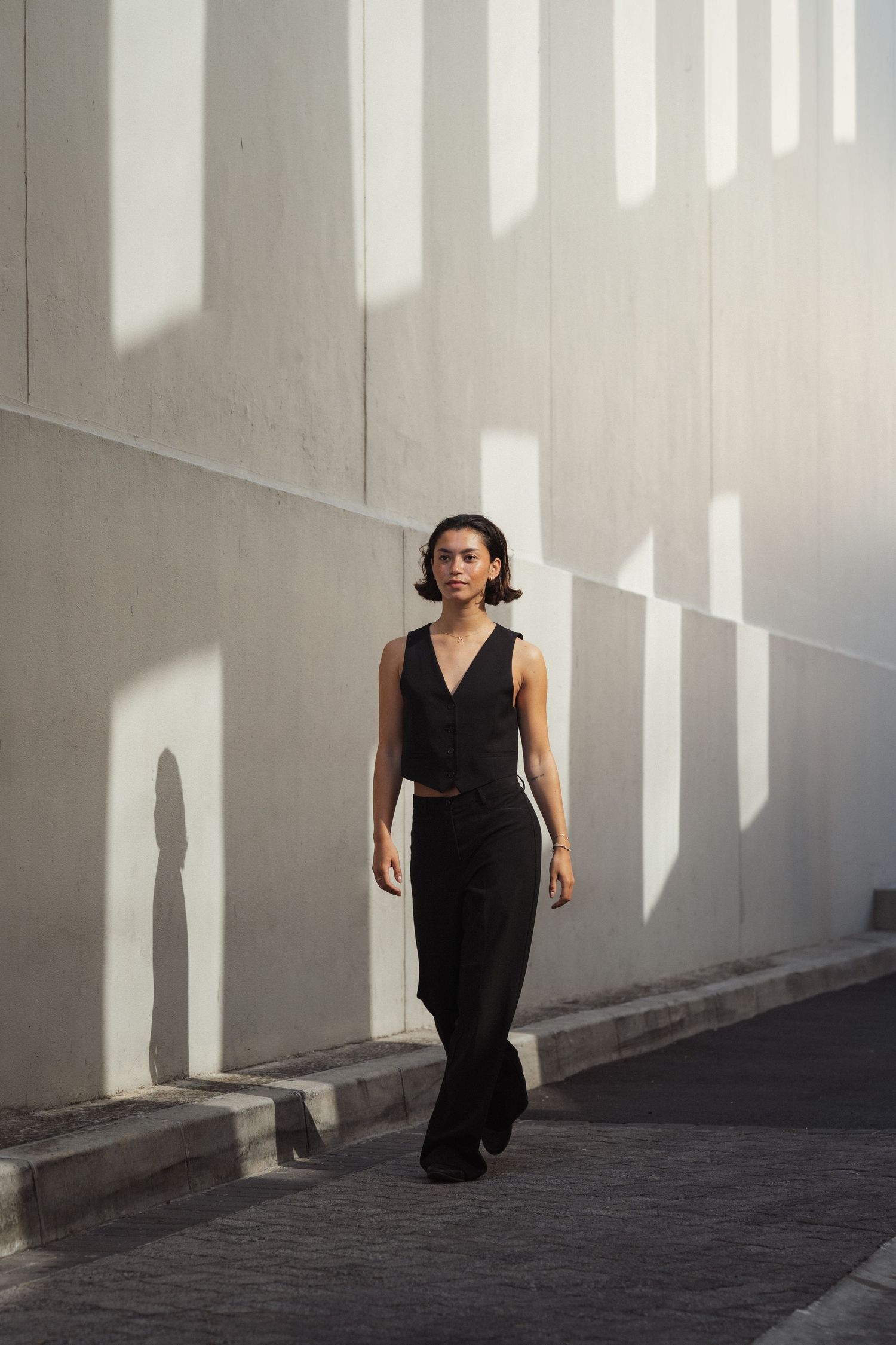 Woman in black crop top and wide-leg pants walking outdoors against a sunlit concrete wall.