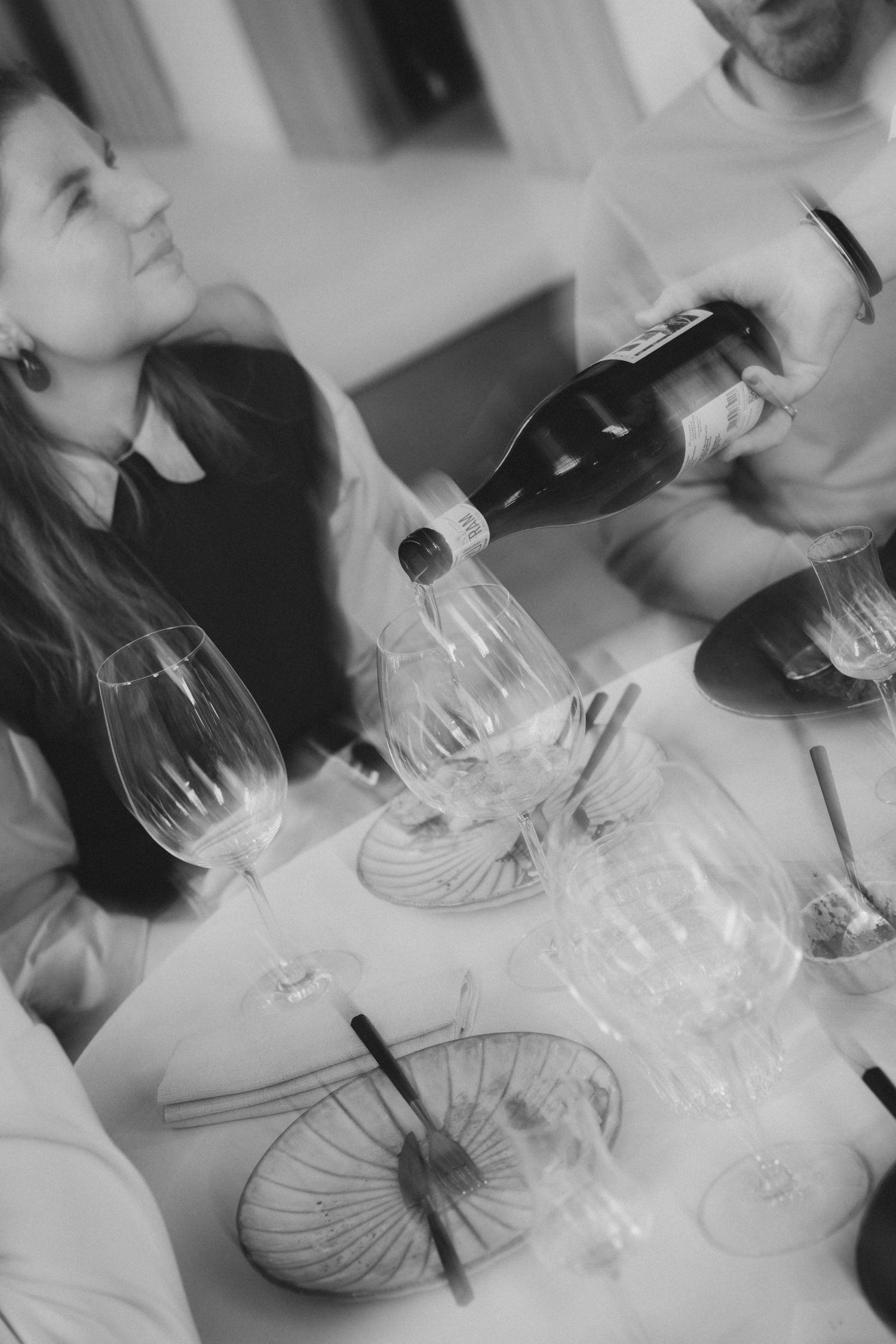 Black and white photo of a woman at a dining table as a man pours wine from a bottle into elegant glasses.