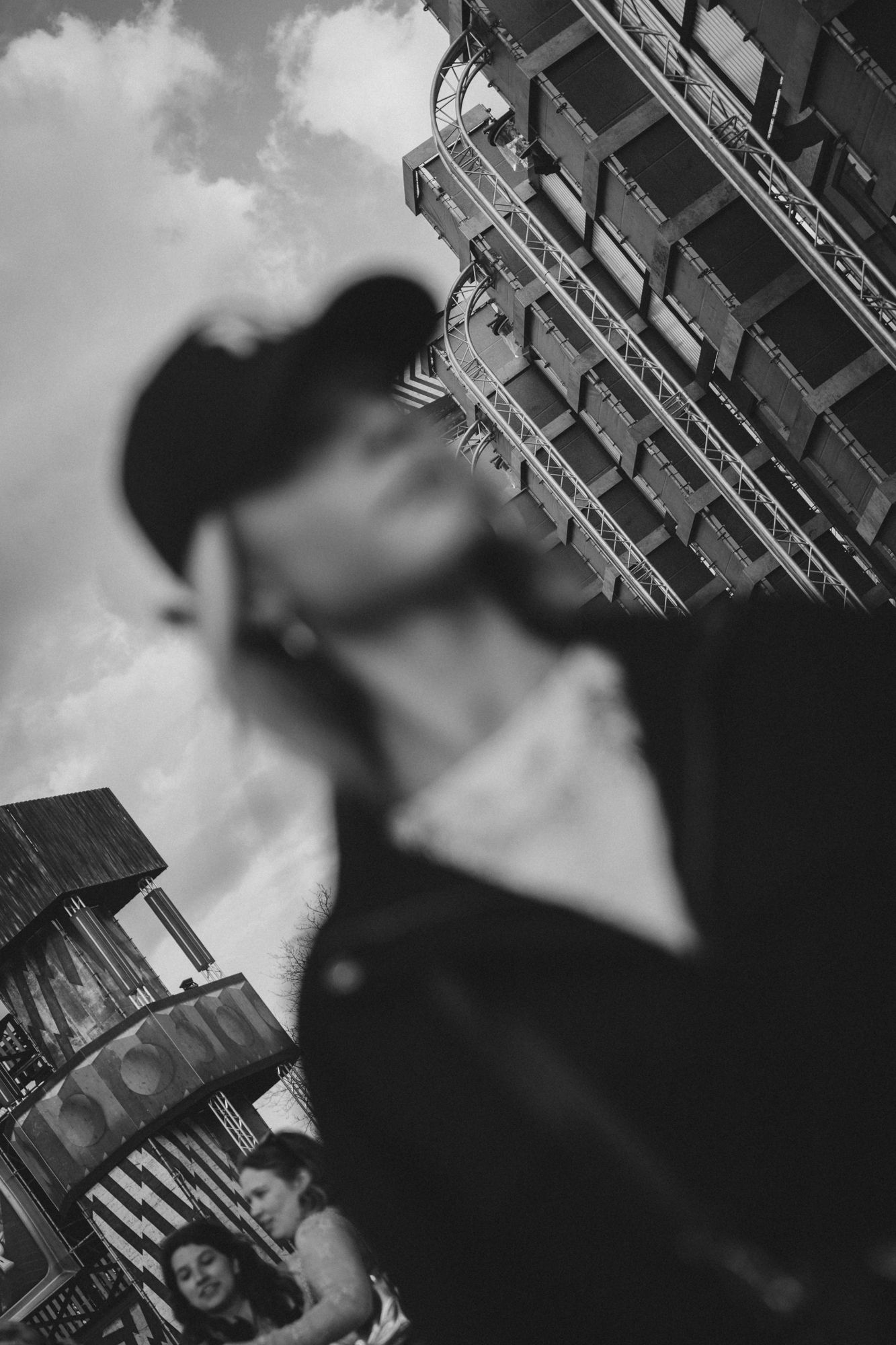 Black and white urban street photo with blurred man in cap, city building and woman visible in background.