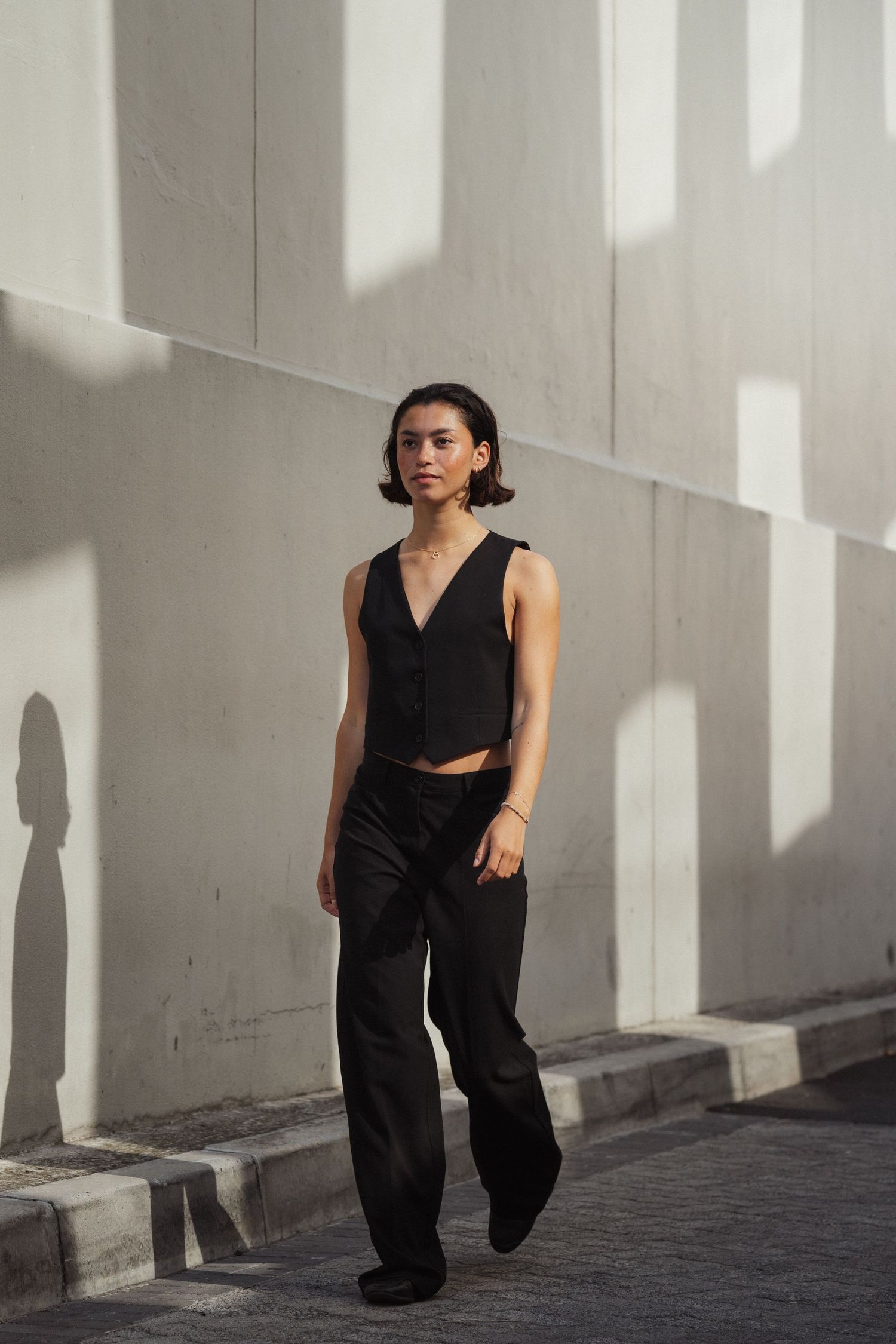 Person in black vest and wide-leg trousers posing against a sunlit white wall outdoors.