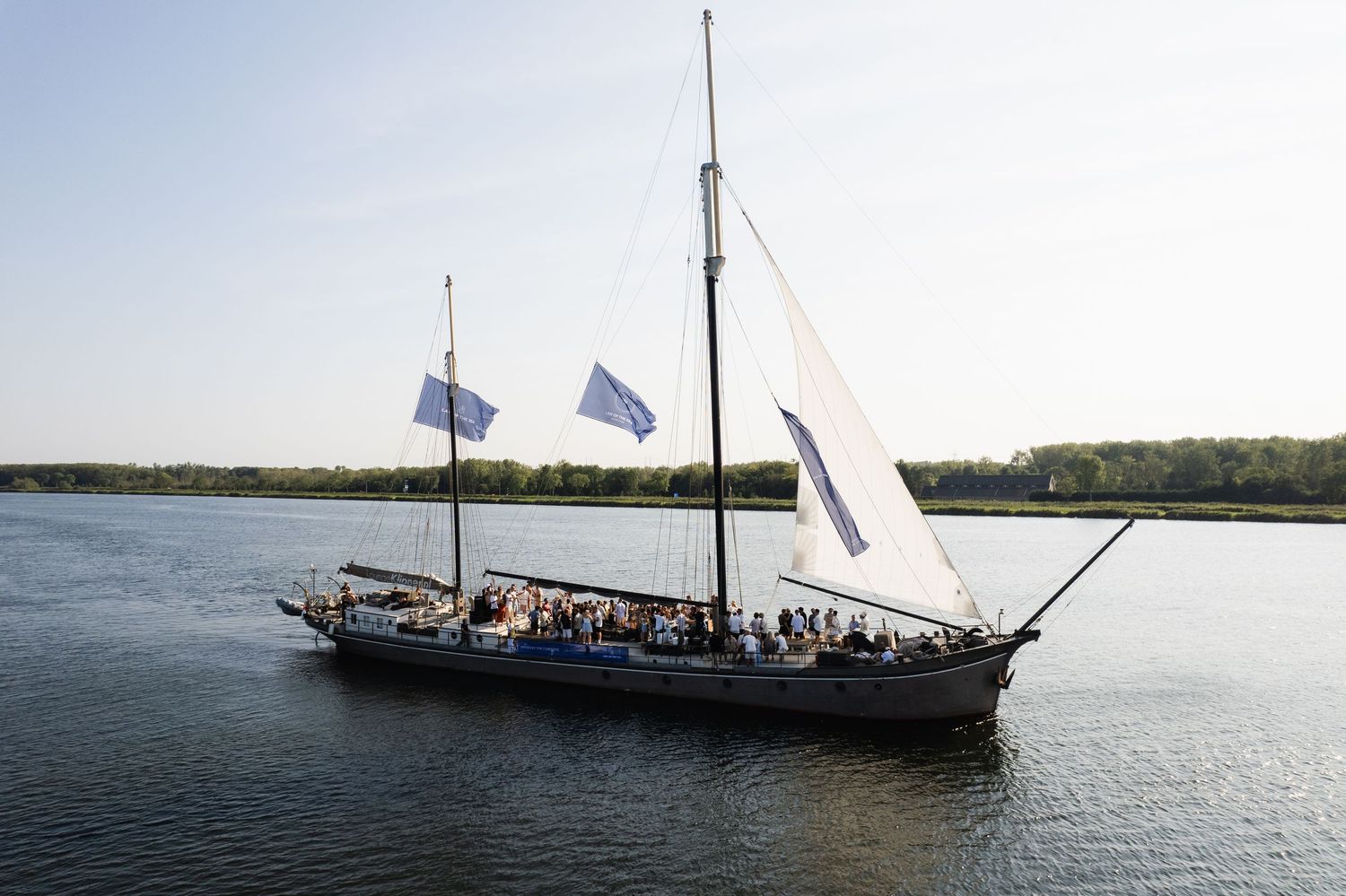 A tall ship with white sails and blue flags cruises along a calm river with passengers on deck.