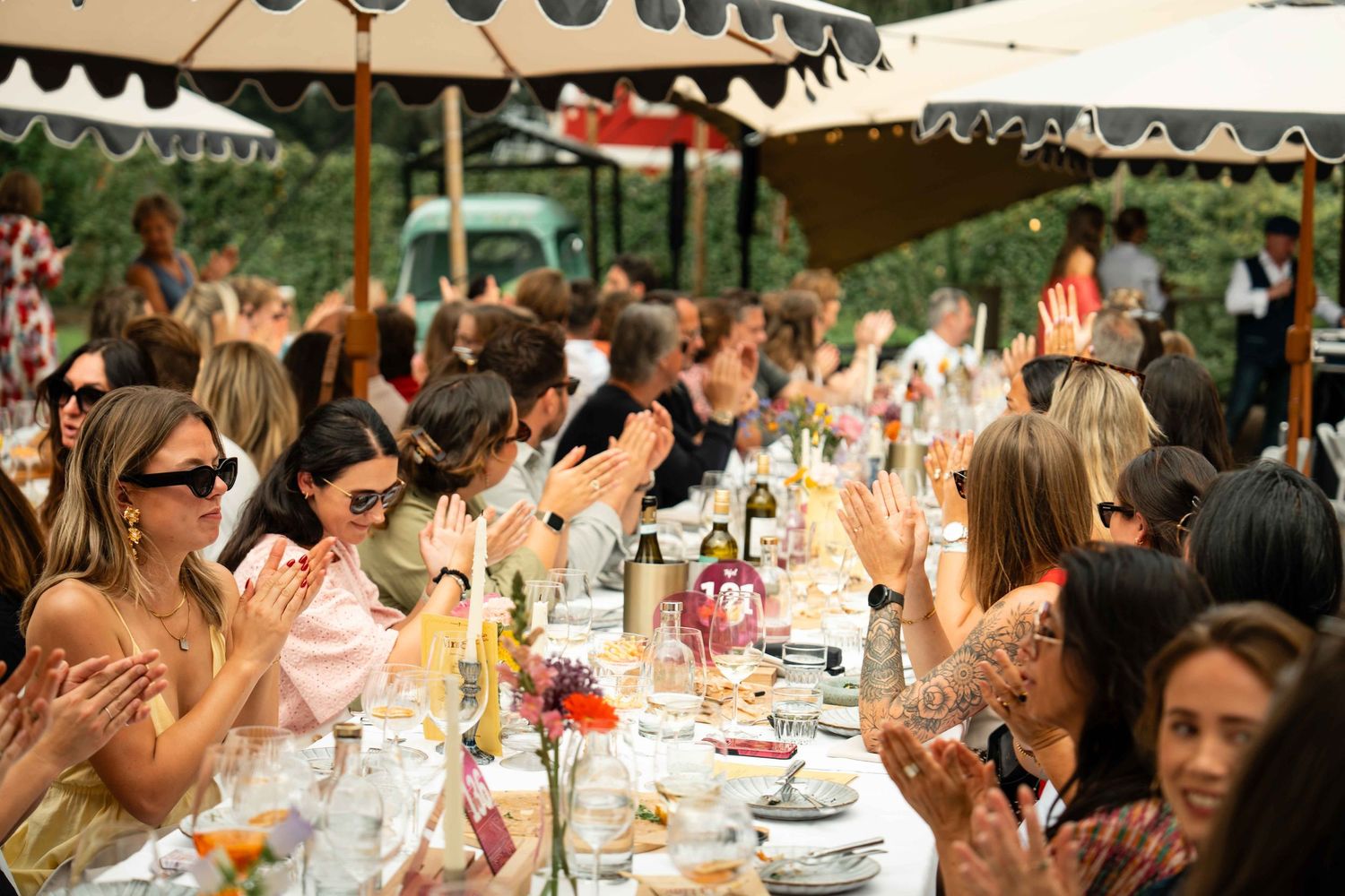Guests clapping at an elegant outdoor garden party under striped marquee tents with long dining tables.