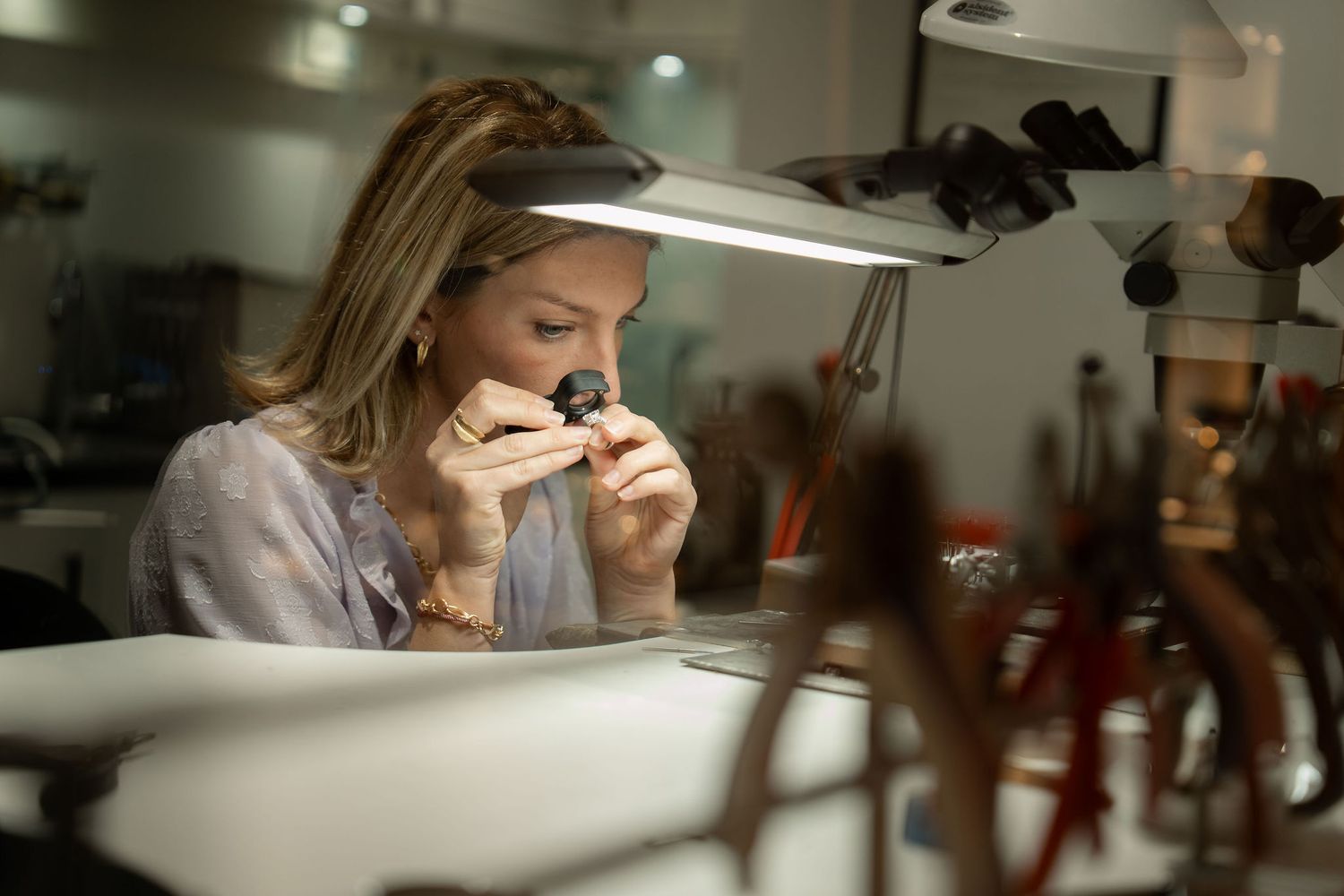 Female jeweler examining a gemstone with a loupe under bright lamp in a professional jewelry workshop.