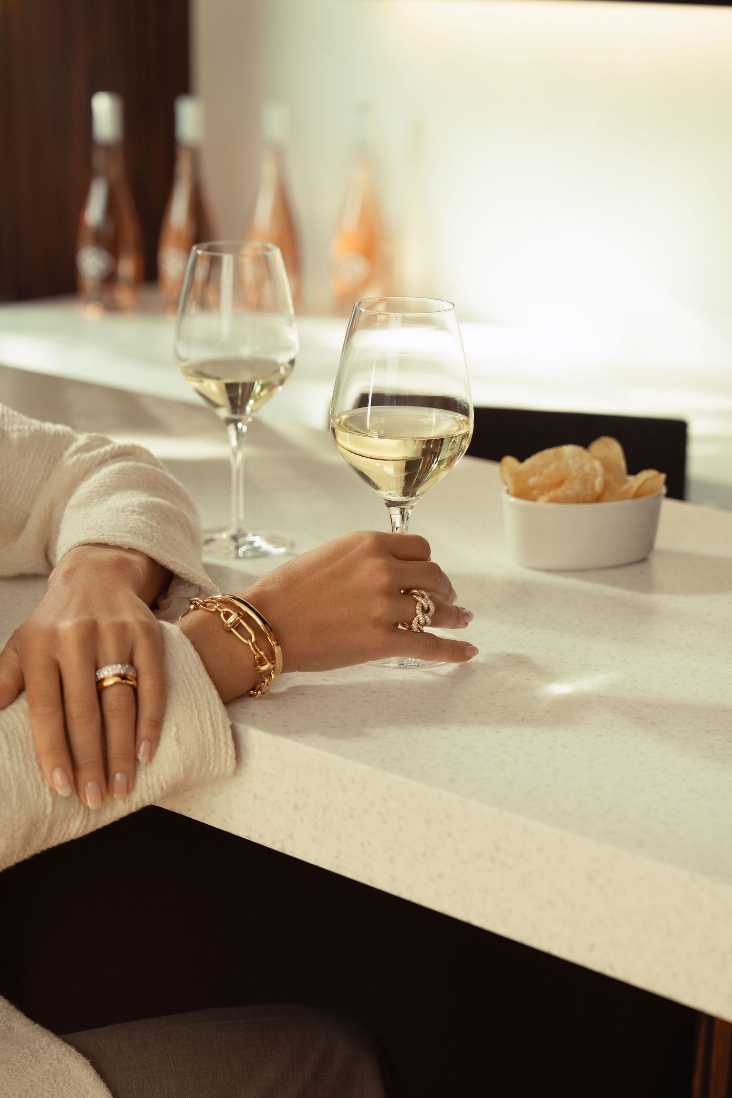 Woman wearing gold bracelets and rings holds a glass of white wine at a marble bar counter.