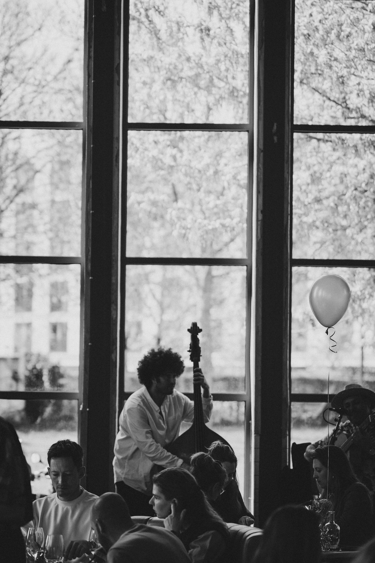 Black and white photo of a musician playing double bass at a crowded indoor event near large windows with a balloon.