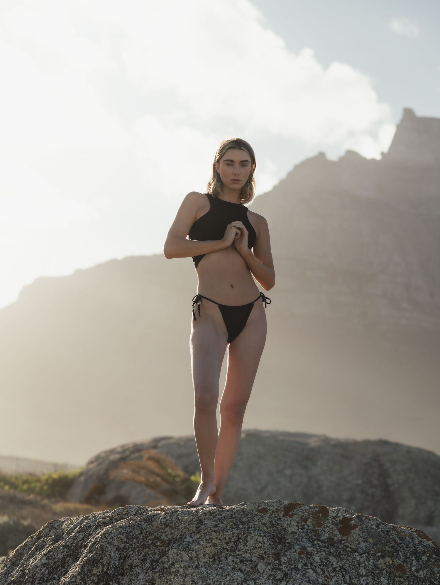 Figure in swimwear posing on rocky terrain with mountains in background.