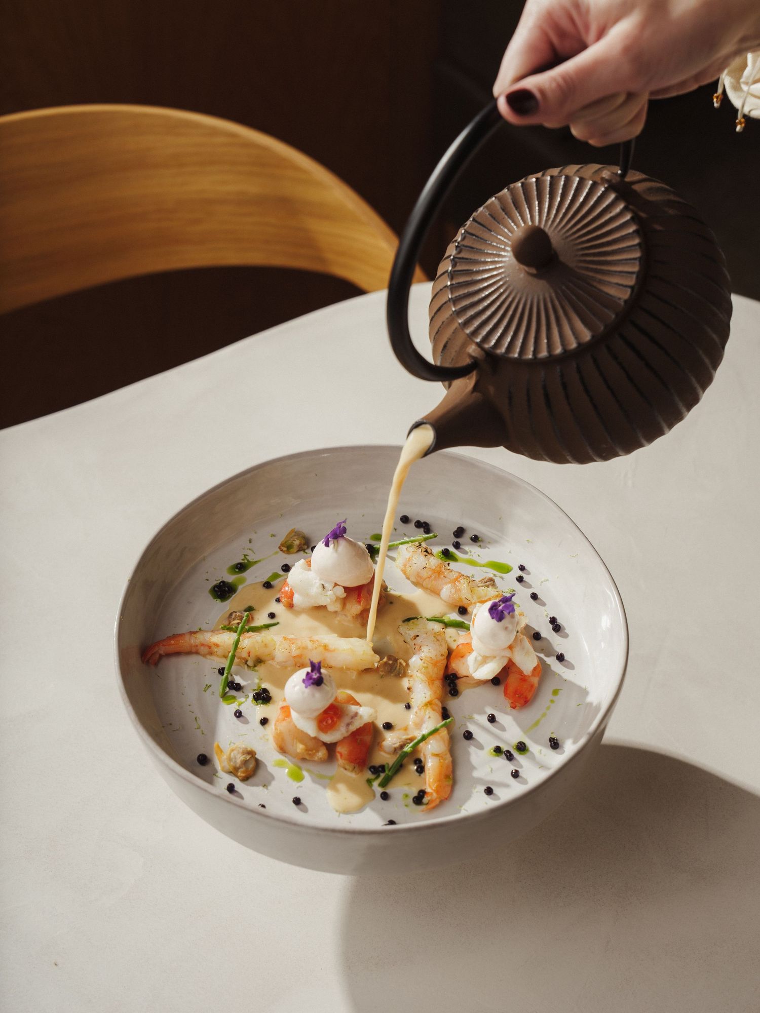 A hand pours creamy broth from a teapot over elegant shrimp dish garnished with herbs and edible flowers in a white bowl.