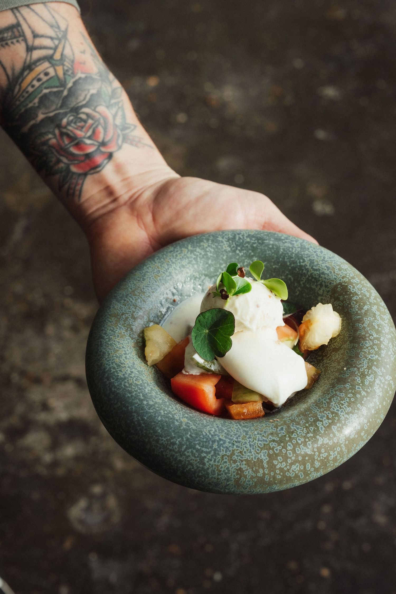 Tattooed hand holding a rustic ceramic bowl with an elegant dish featuring white fish, microgreens, and colorful vegetables.