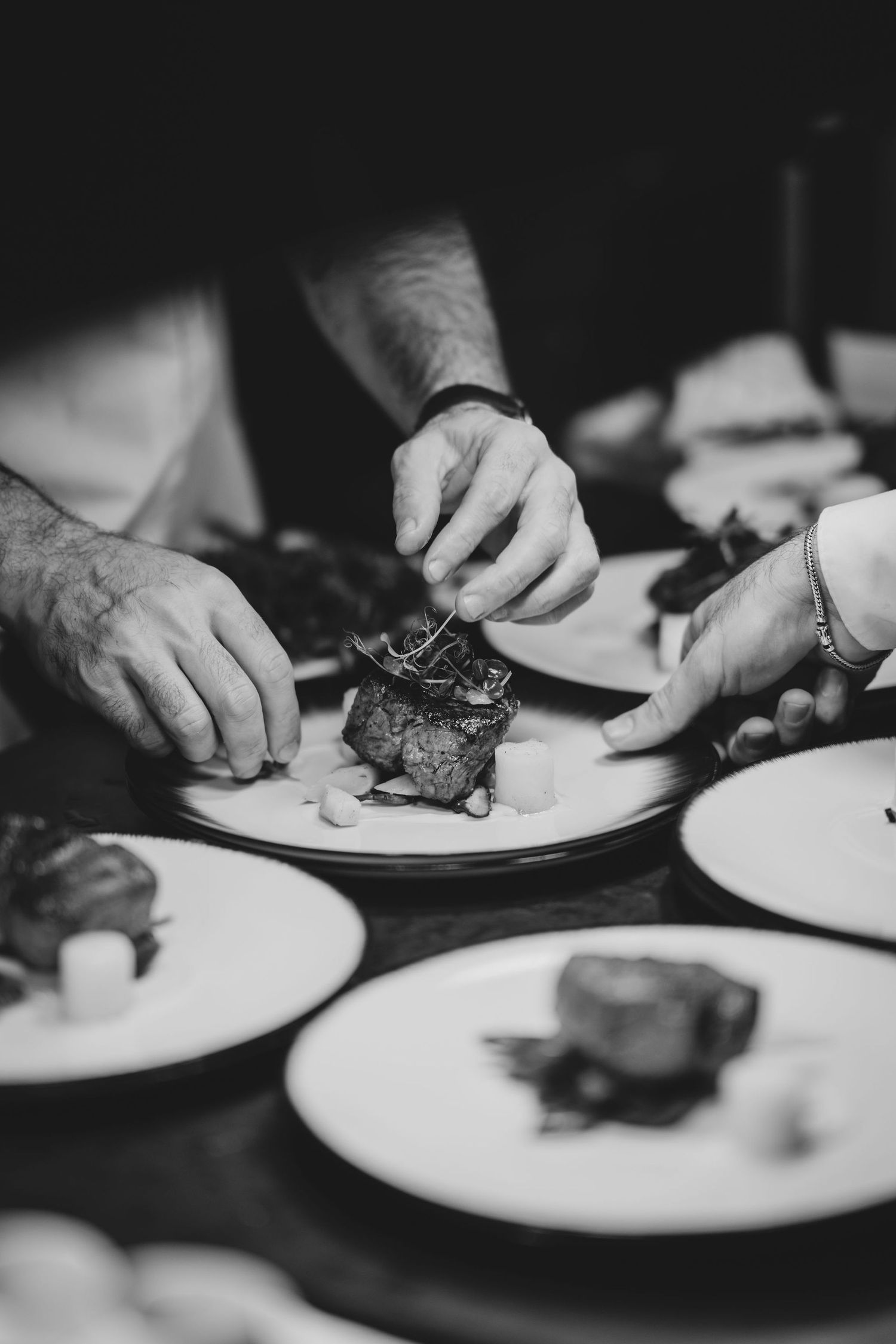 Black and white photo of a chef carefully plating gourmet dishes on white plates in a professional kitchen.