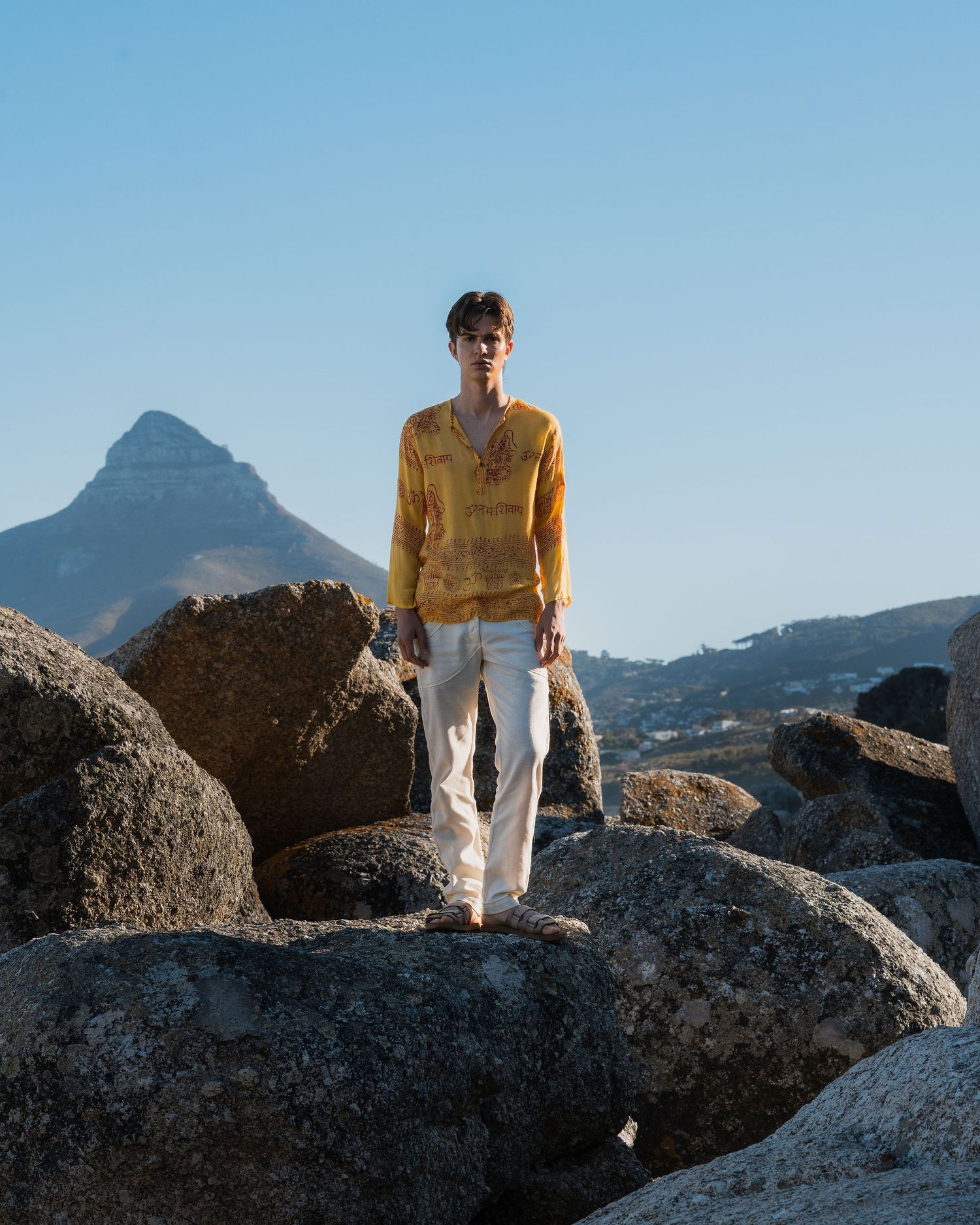 Man in yellow shirt and white pants standing on rocks with mountain backdrop under clear blue sky.