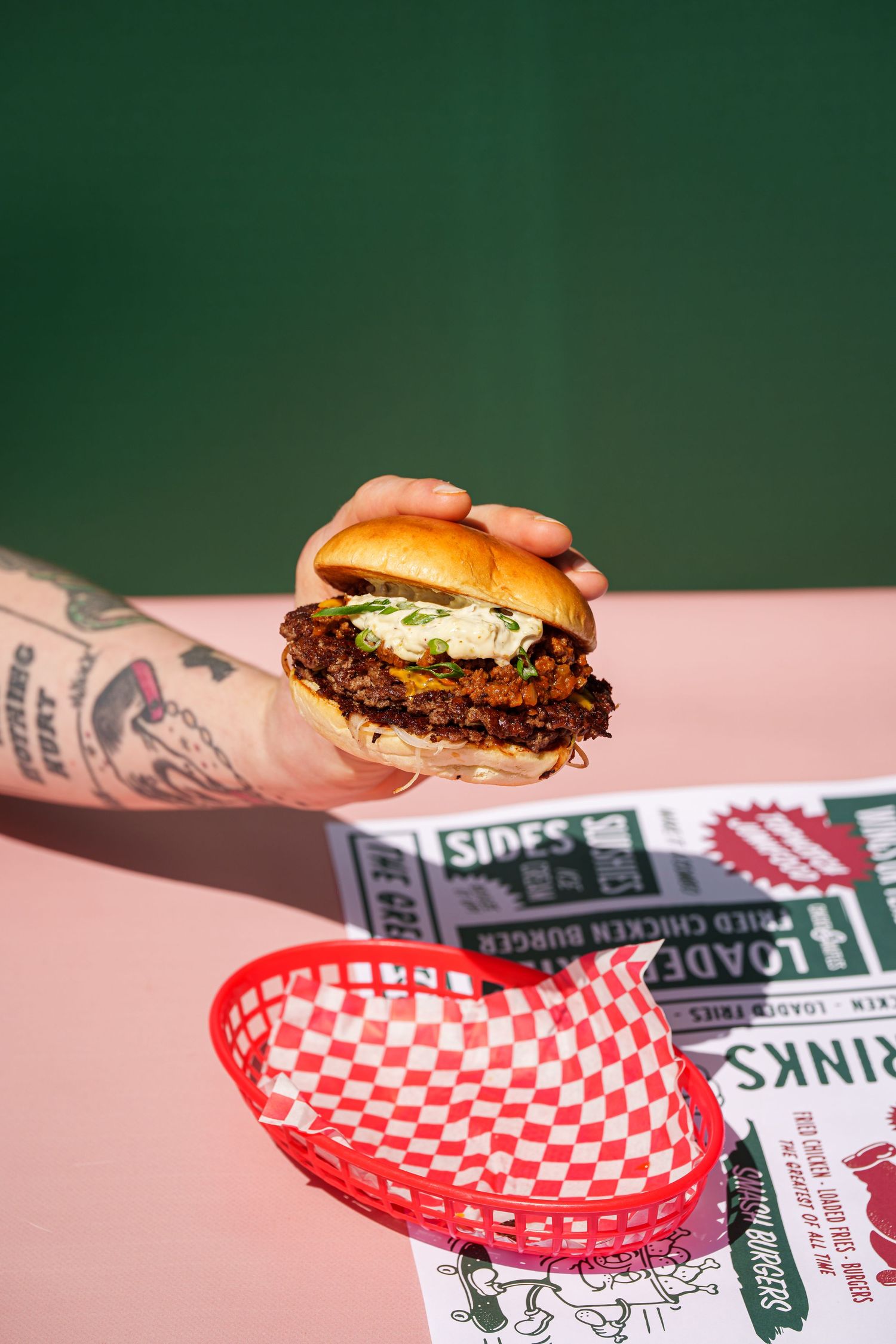 Tattooed hand holding a smashed burger with toppings above a red checkered basket and menu on pink surface.
