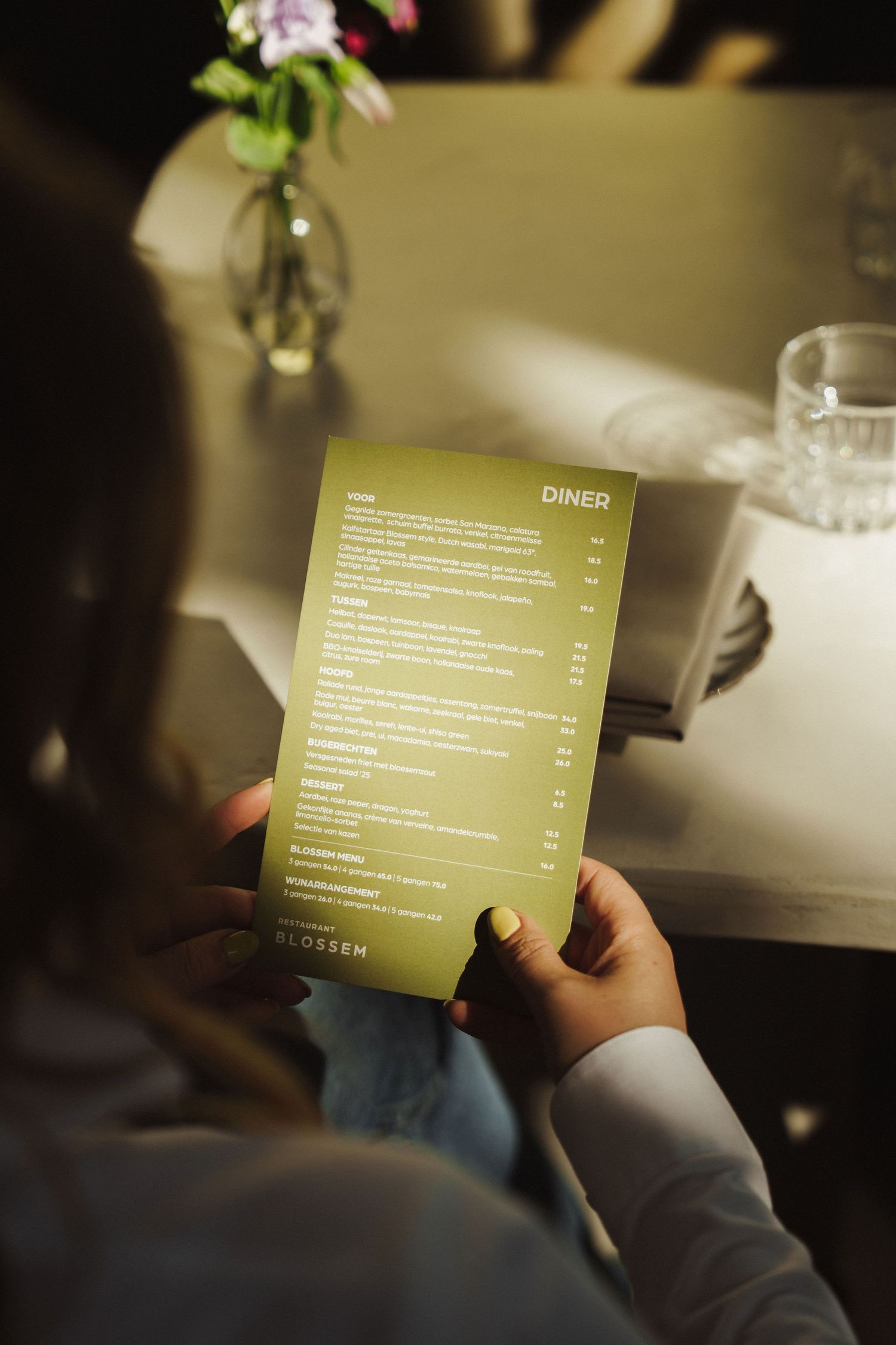 Person holding a green diner menu at a restaurant table with a floral vase and glass in the background.