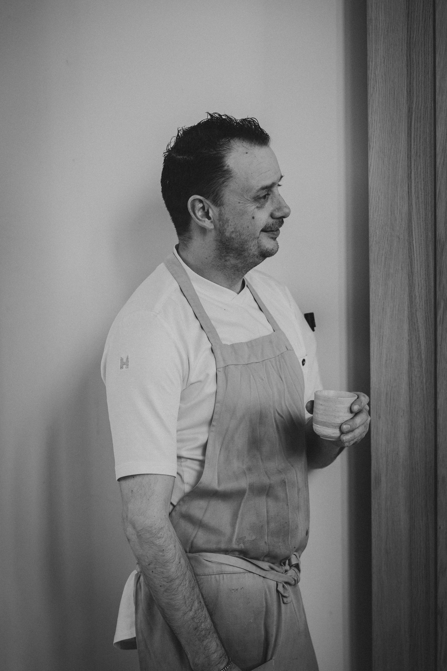 Black and white photo of a male chef in apron holding a coffee cup, standing thoughtfully against a wall.