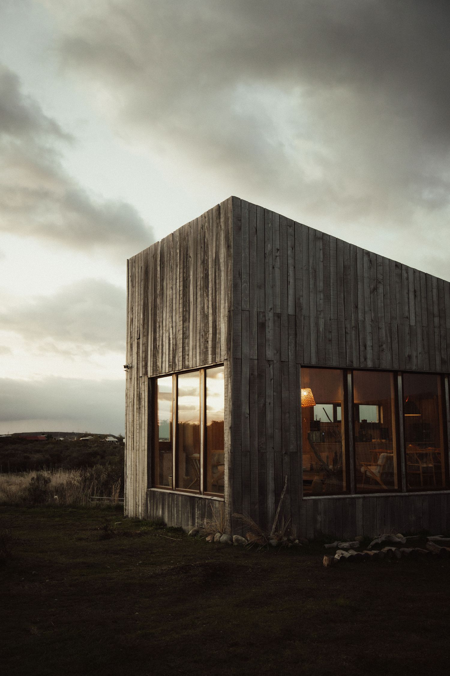Rustic wooden cabin with large glass windows reflecting a golden sunset under dramatic cloudy skies in a remote landscape.