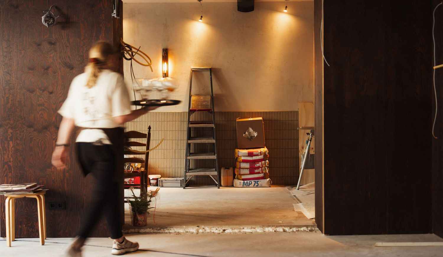 A blurred waiter carries a tray past a room under renovation with a ladder and construction materials.