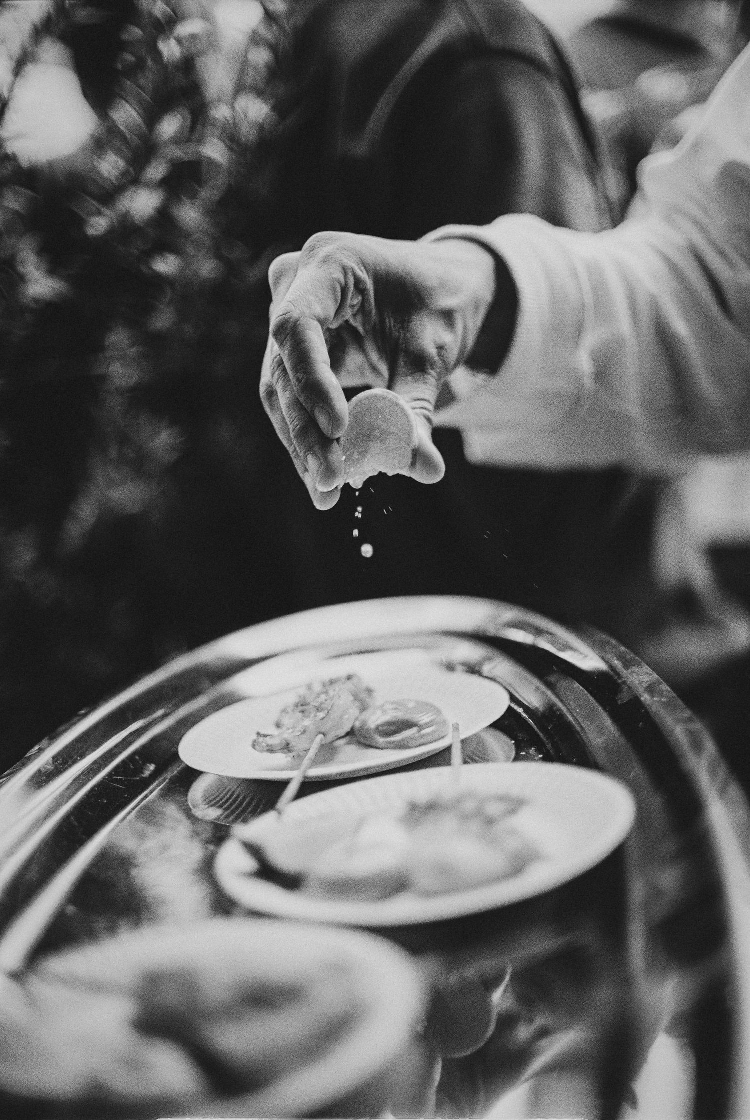 A waiter seasons a gourmet dish under a silver cloche in a black and white fine dining scene.