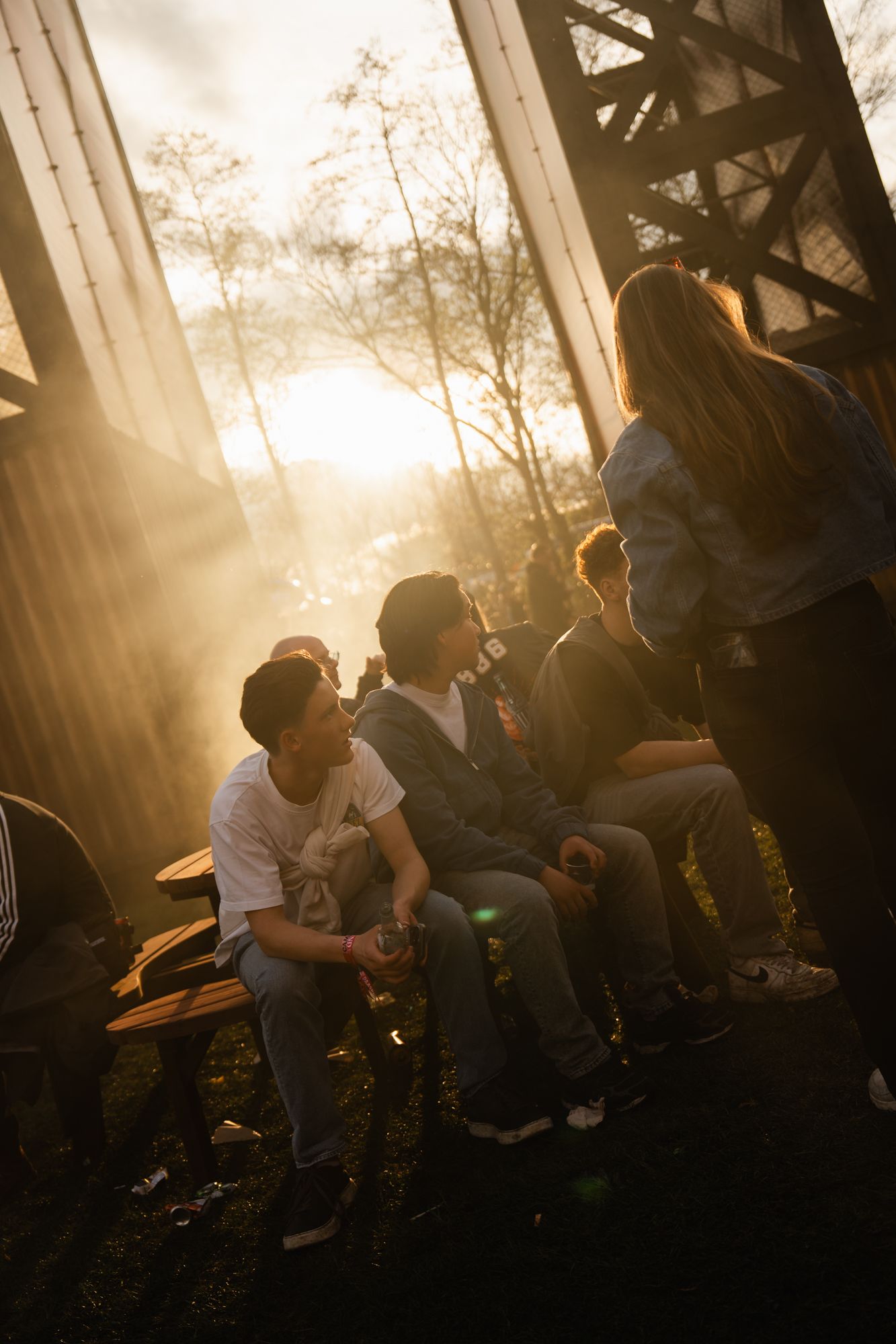 Group of young people socializing on a bench outdoors at golden sunset hour near an industrial structure.