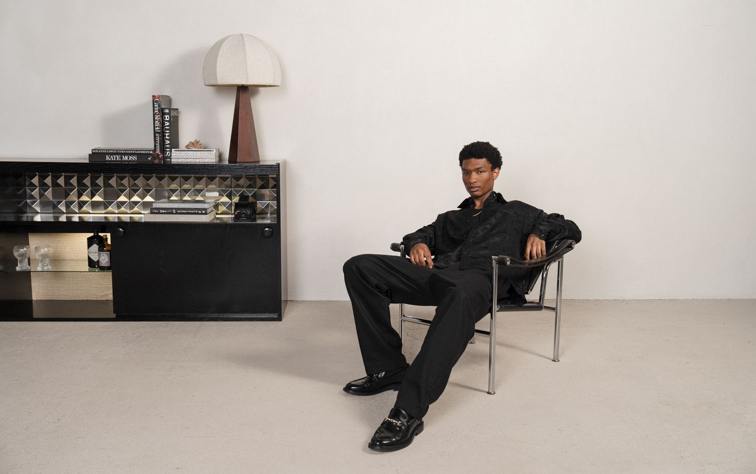 A person sits in a modern armchair against a white wall with a minimalist sideboard and lamp in the background.