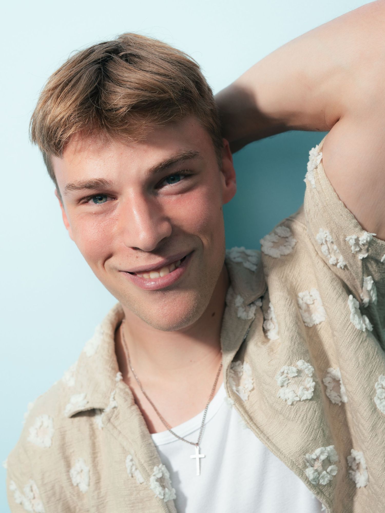 Young man with short blonde hair smiling, wearing a beige floral shirt and cross necklace against a light blue background.
