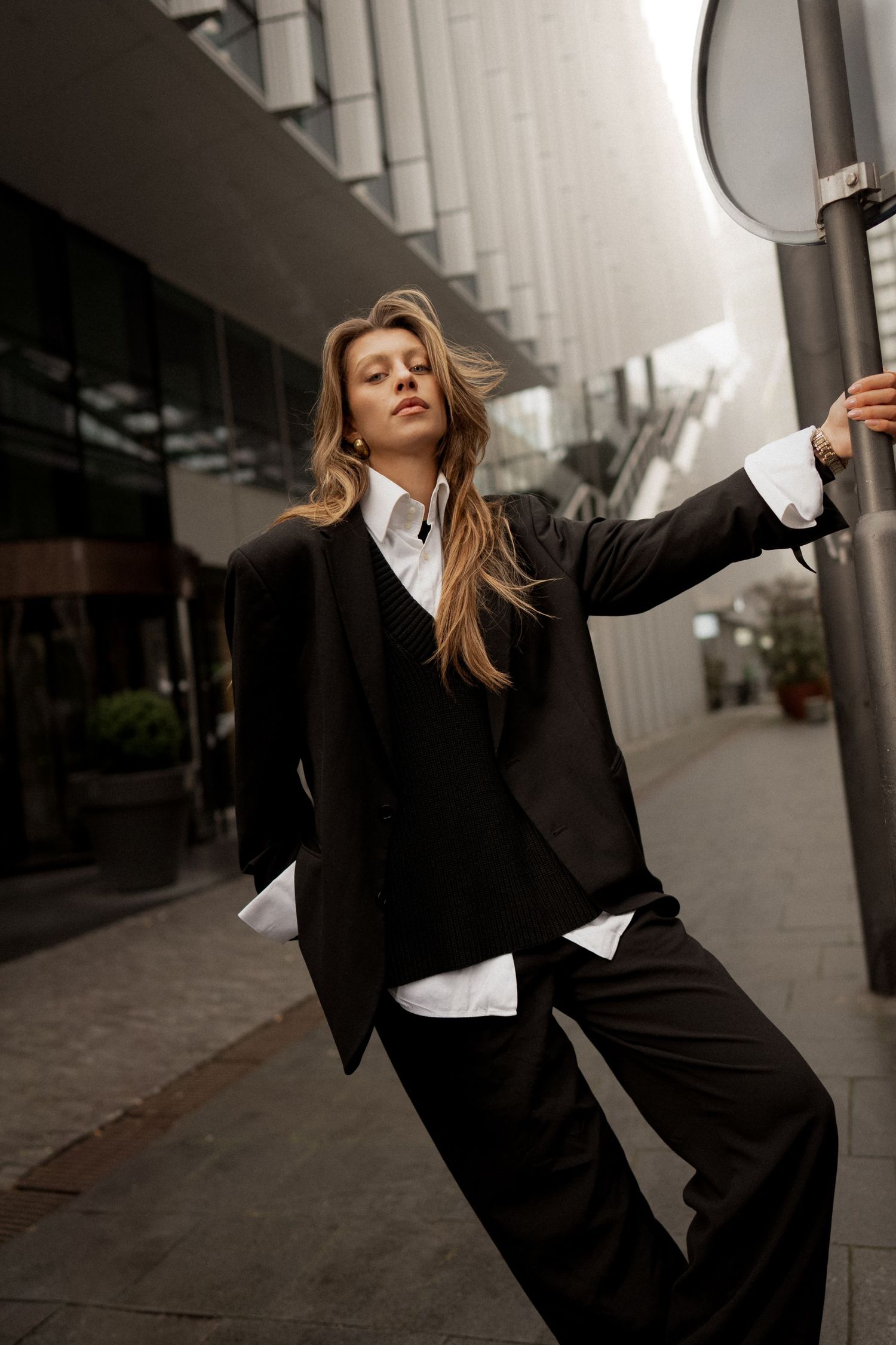 Woman in black oversized suit and white shirt leans against a pole on a city street.