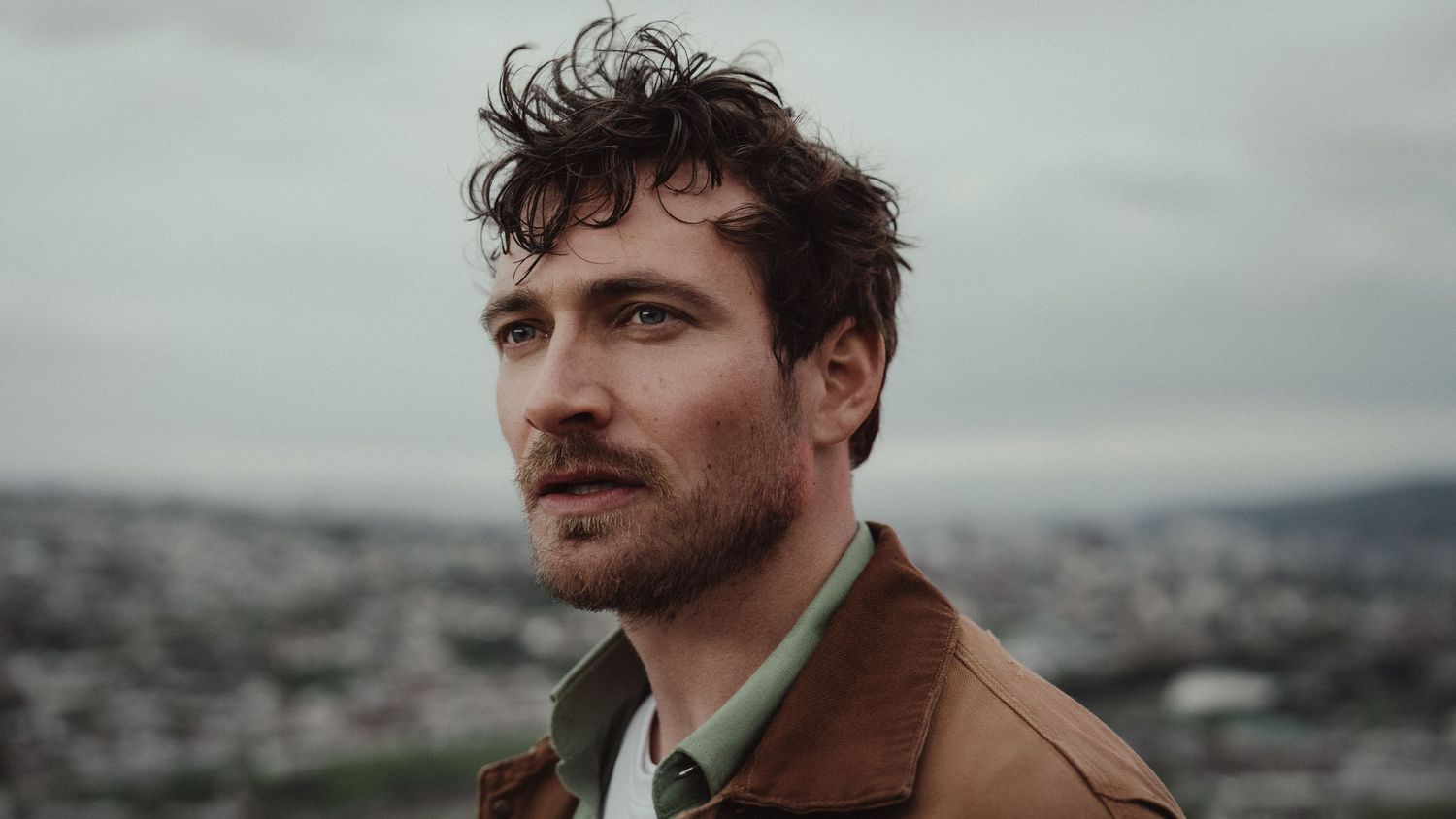 Young man with curly hair in brown jacket gazes thoughtfully against a cloudy city skyline backdrop.