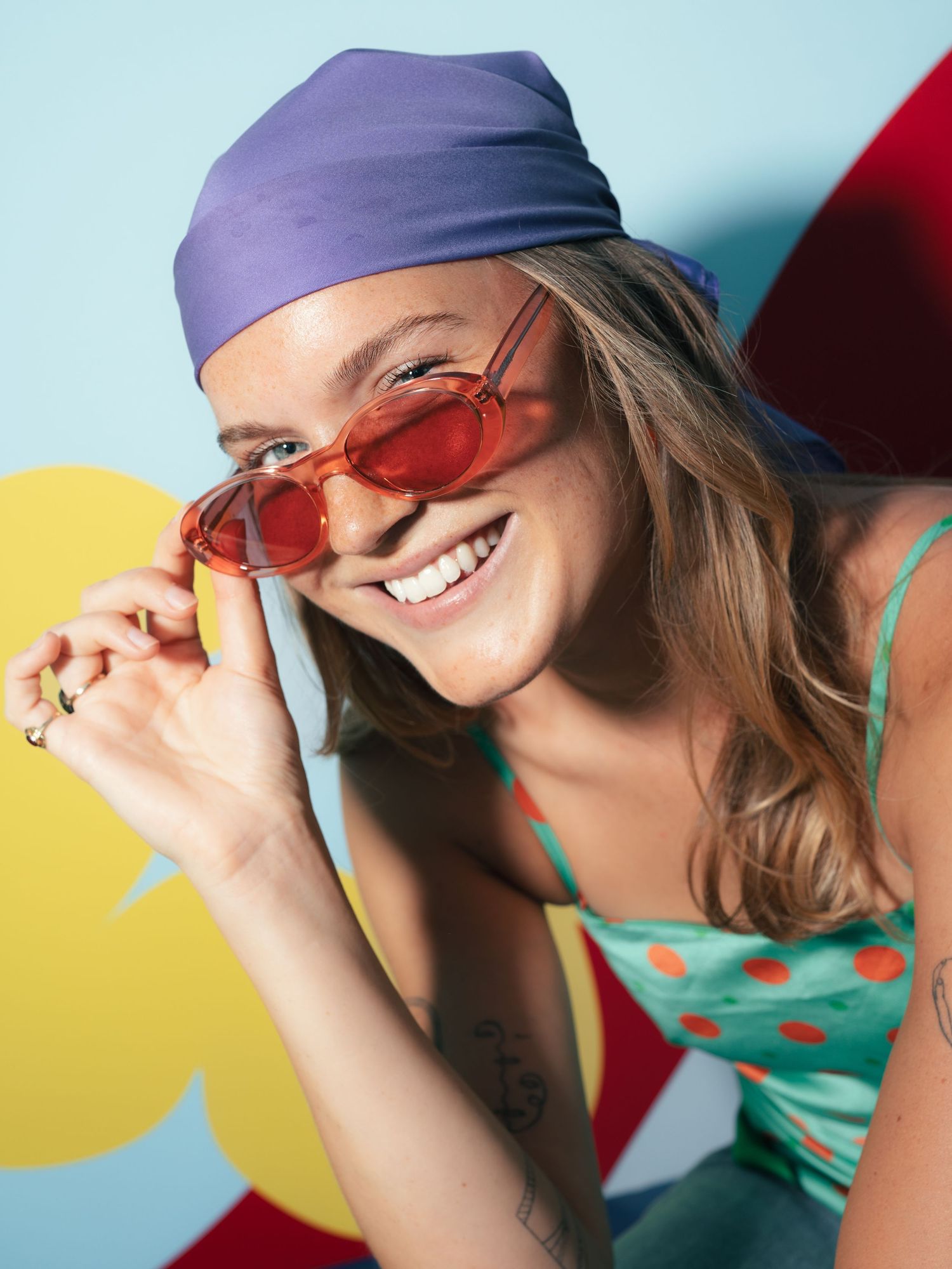 Smiling woman in purple bandana and red oval sunglasses wearing a teal polka dot top in a colorful setting.