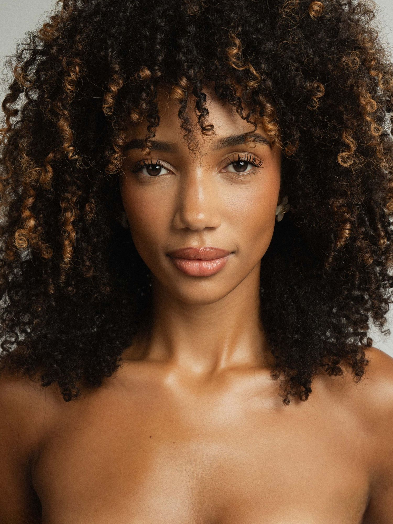 A young woman with voluminous curly black hair with caramel highlights poses against a light gray background.