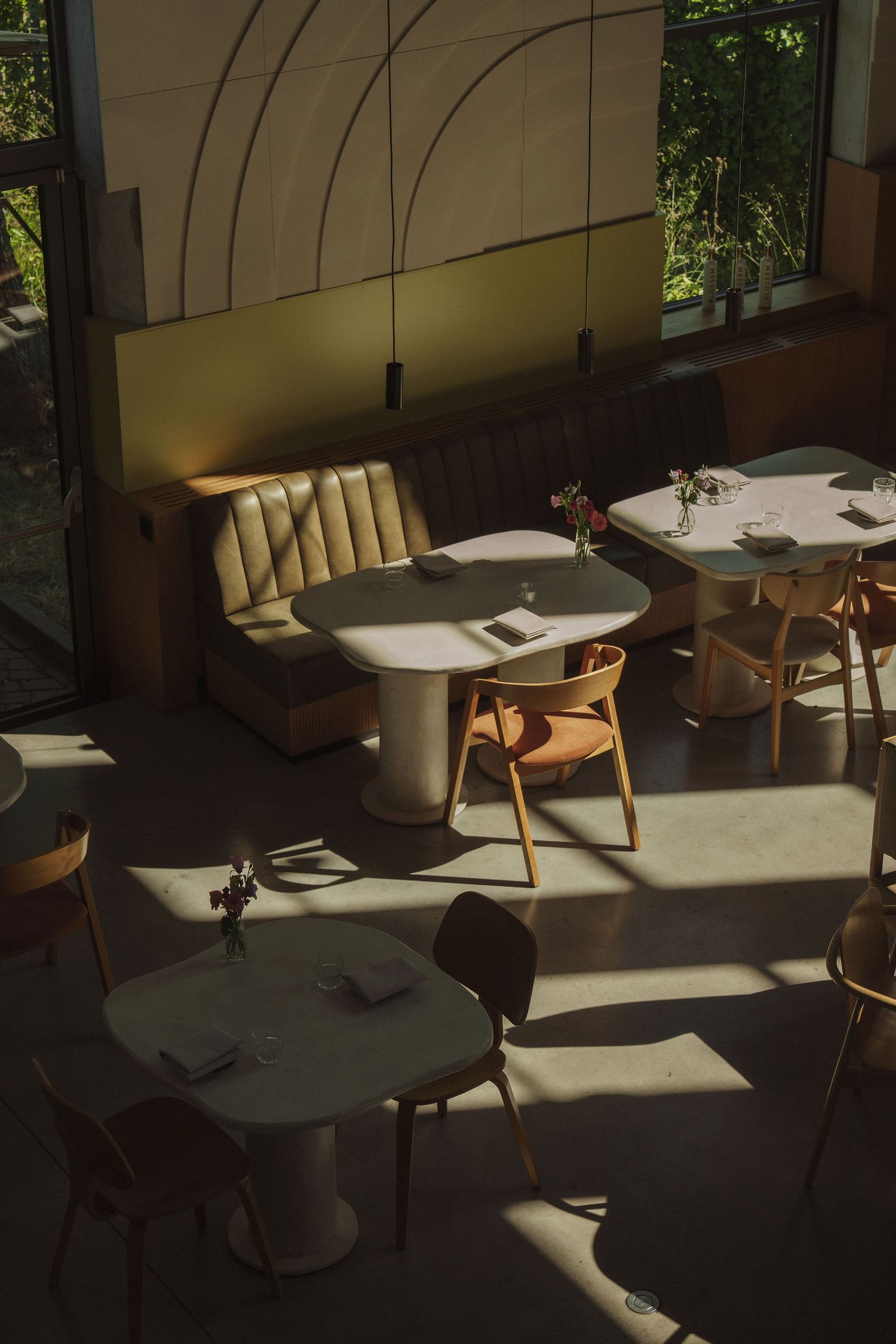Sunlit modern restaurant interior with round white tables, wooden chairs, and a beige booth seat casting warm shadows.