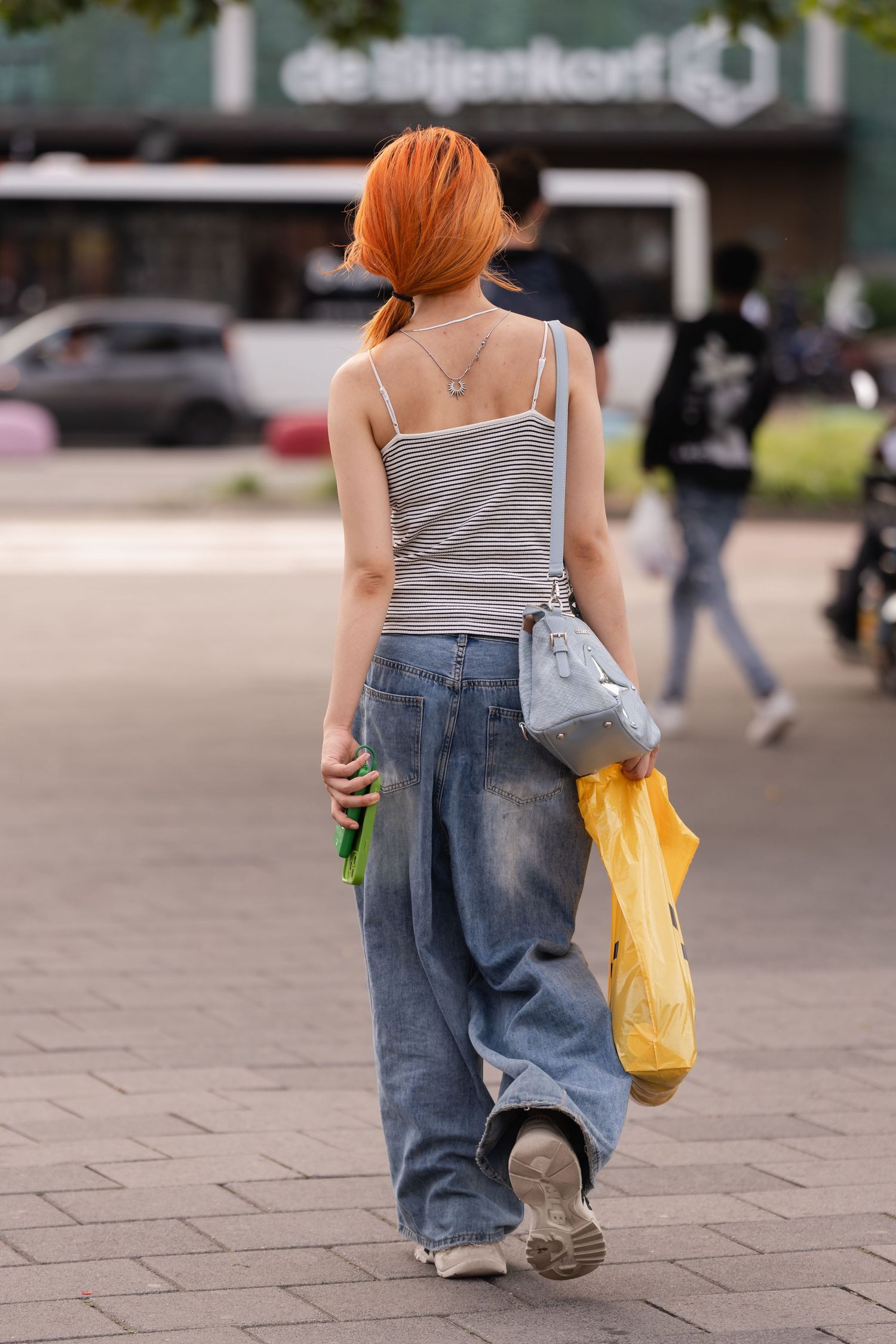 Woman with red hair in striped tank top and baggy jeans carrying bags on a city square.