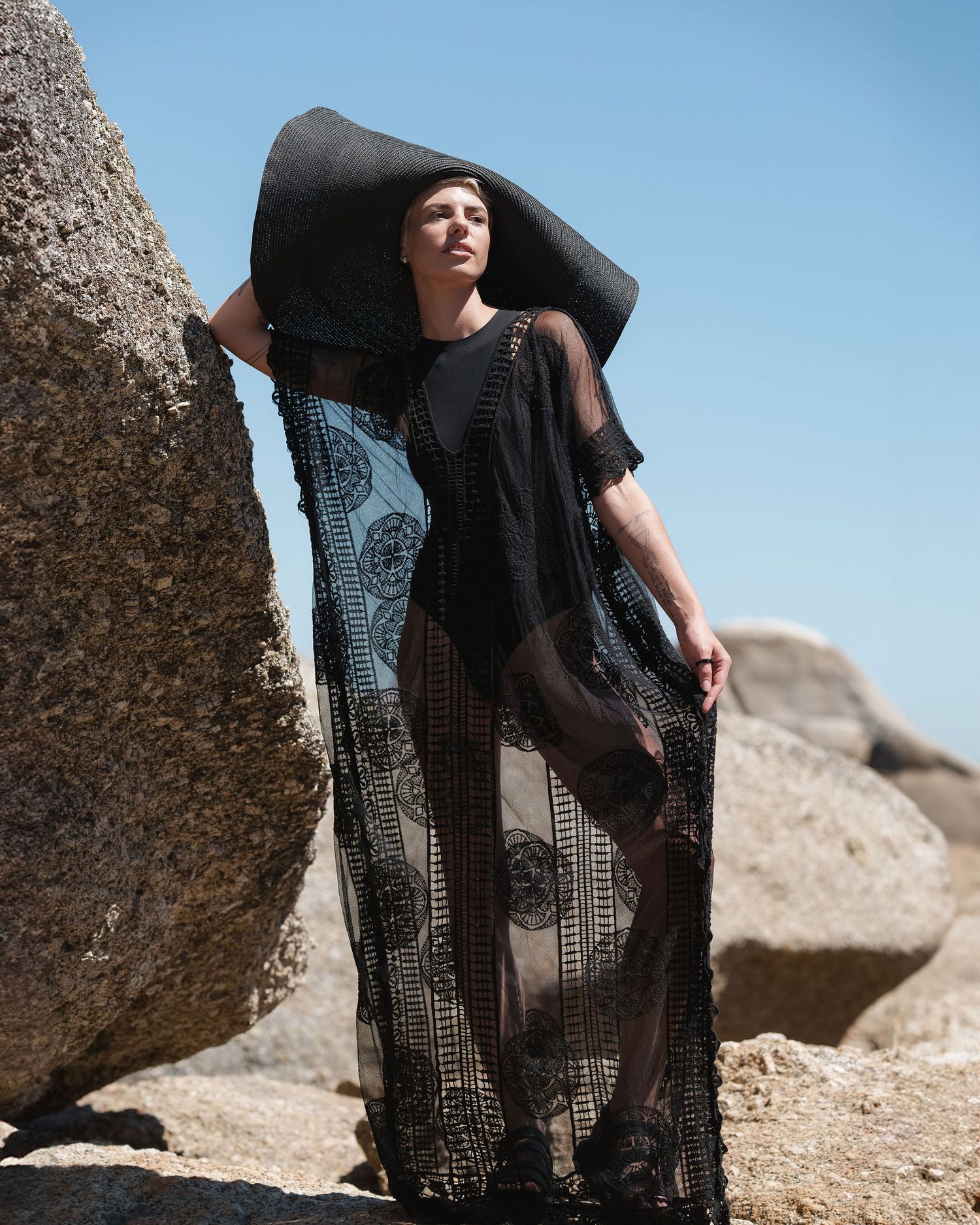 Model wearing a sheer black lace maxi kimono and wide-brim hat, posing against large rocks under a clear blue sky.