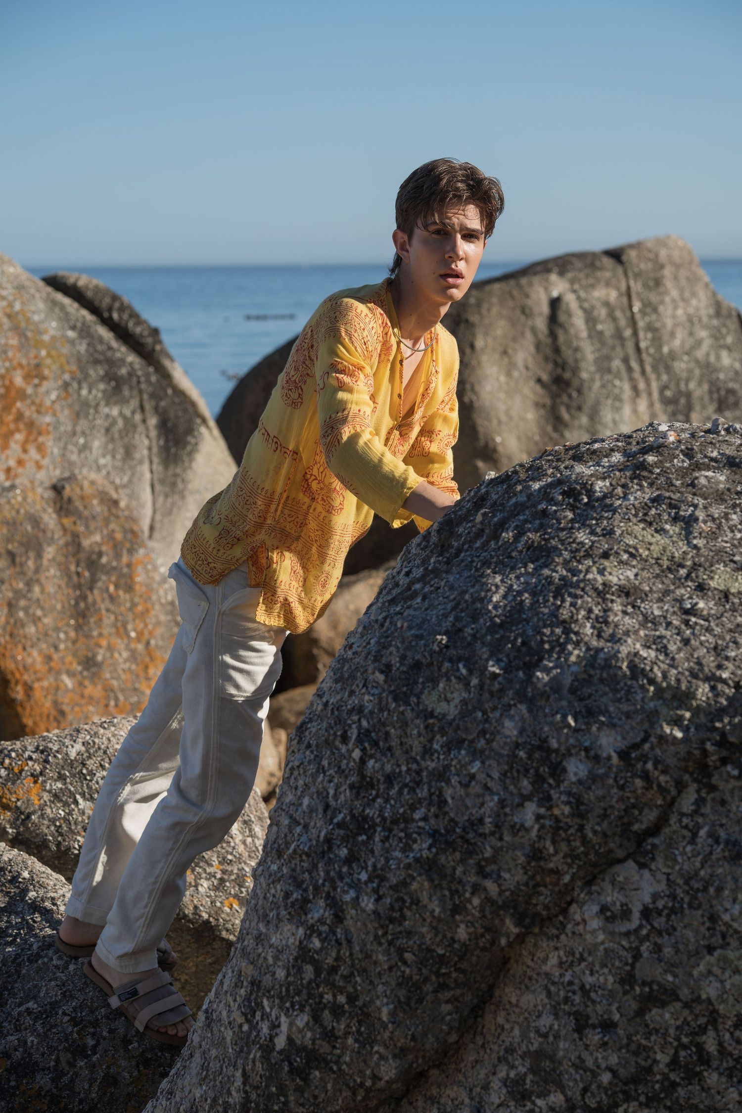 Young man in yellow patterned shirt and white pants climbing rocky boulders by the ocean on a sunny day.