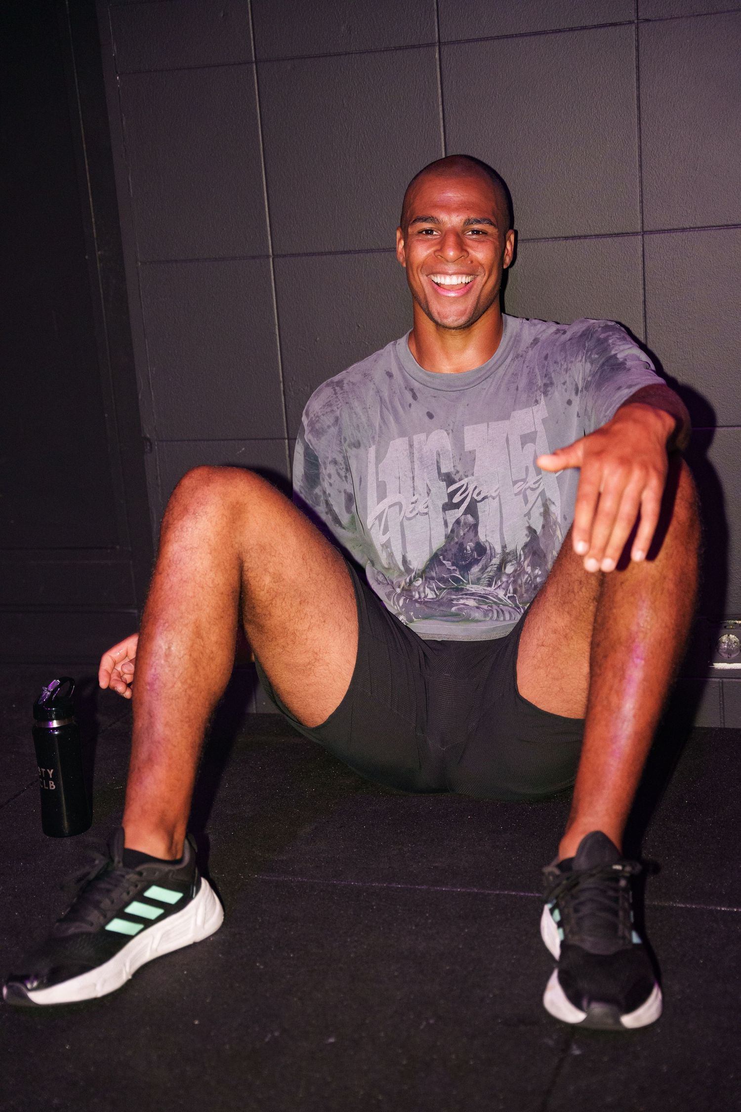 Smiling athletic man sitting on gym floor wearing gray t-shirt, black shorts, and Adidas sneakers with water bottle.