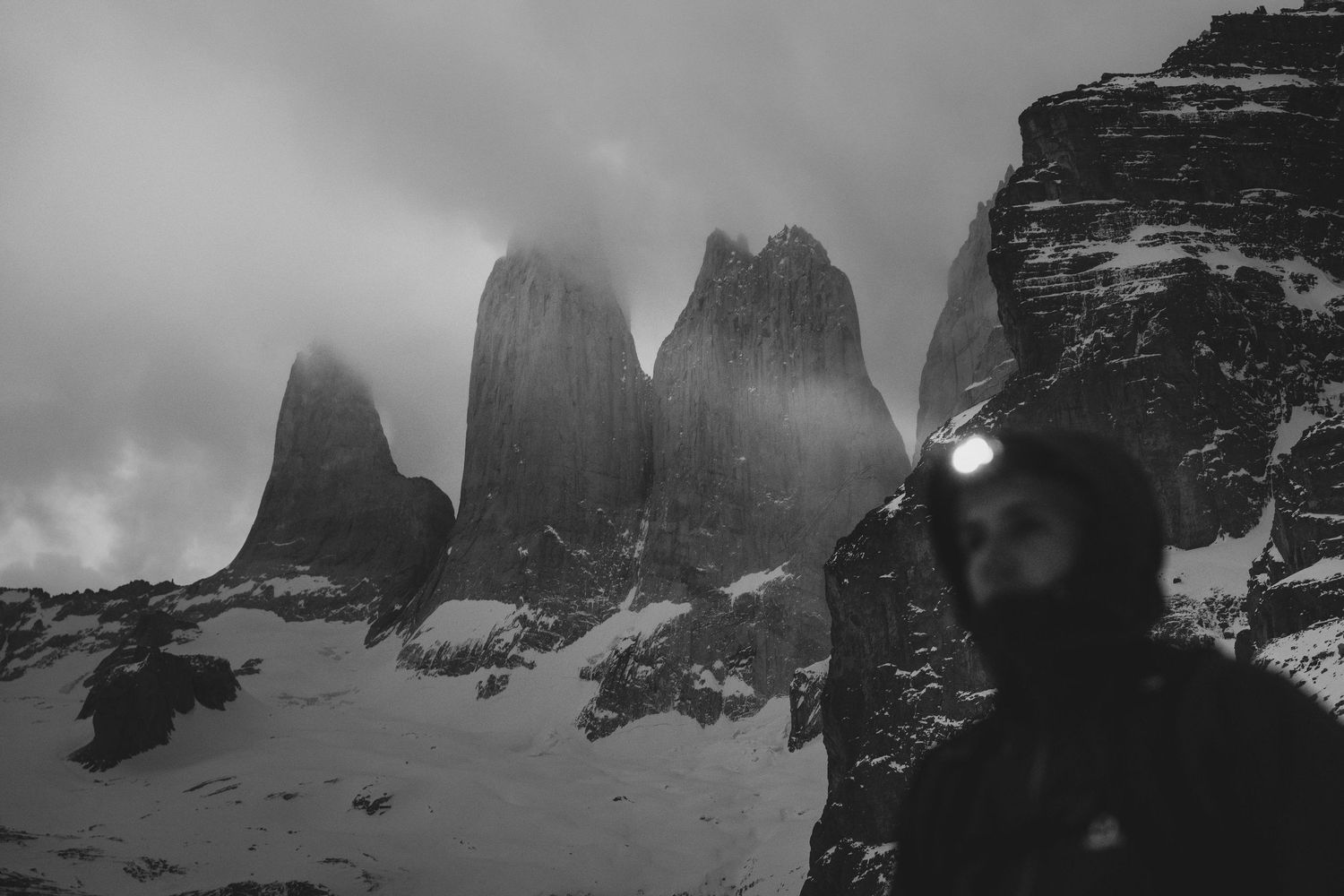 Black and white photo of a hiker with headlamp before Torres del Paine towers in snowy Patagonia.