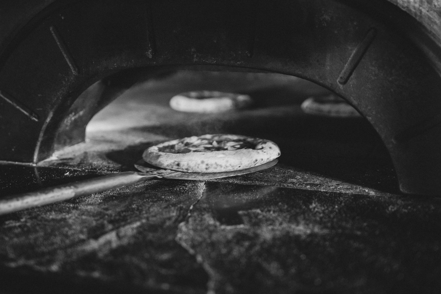 Black and white photo of pizzas baking inside a traditional wood-fired stone oven.