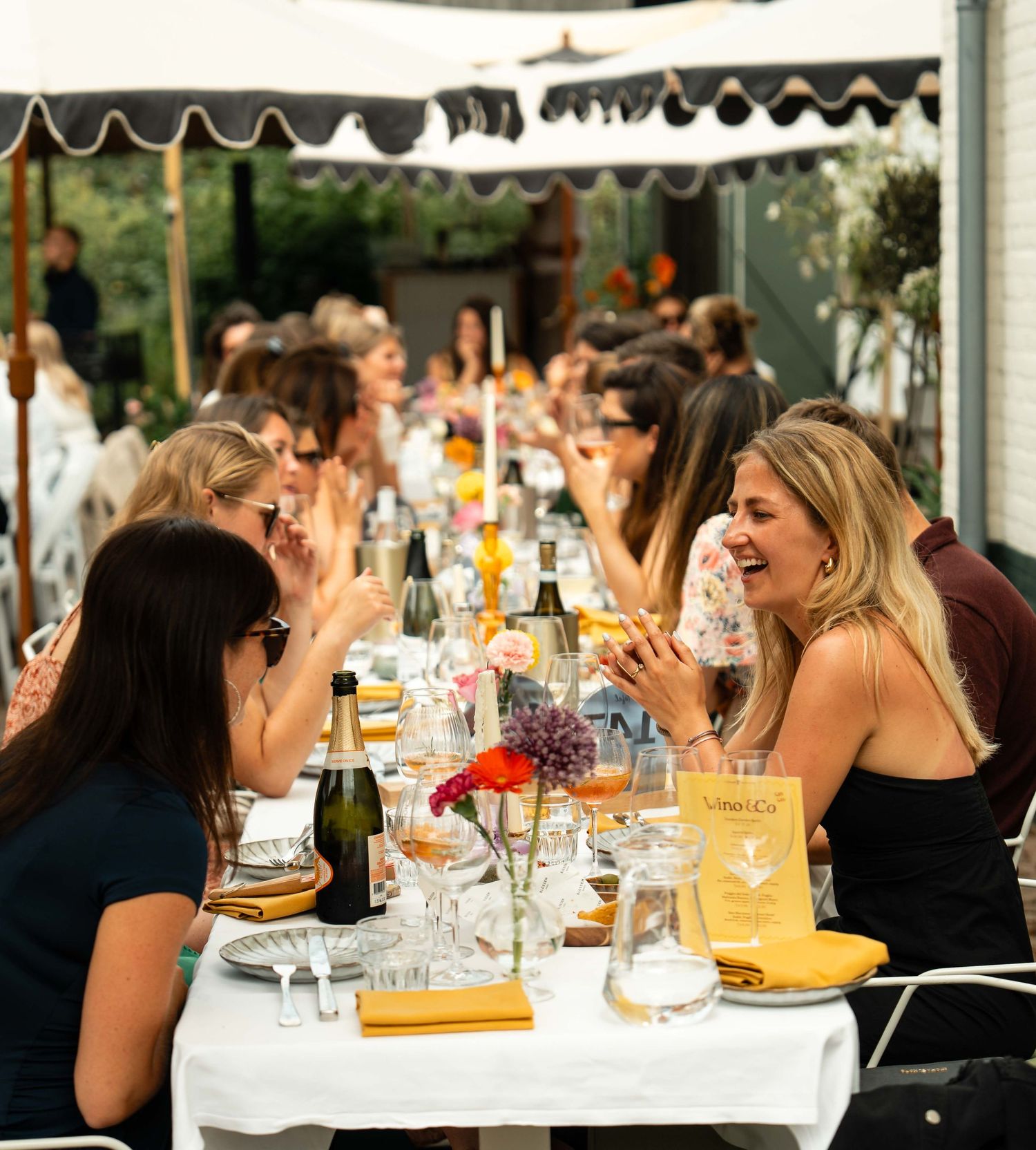 Women laughing and socializing at an elegant outdoor dining event with champagne and floral centerpieces.