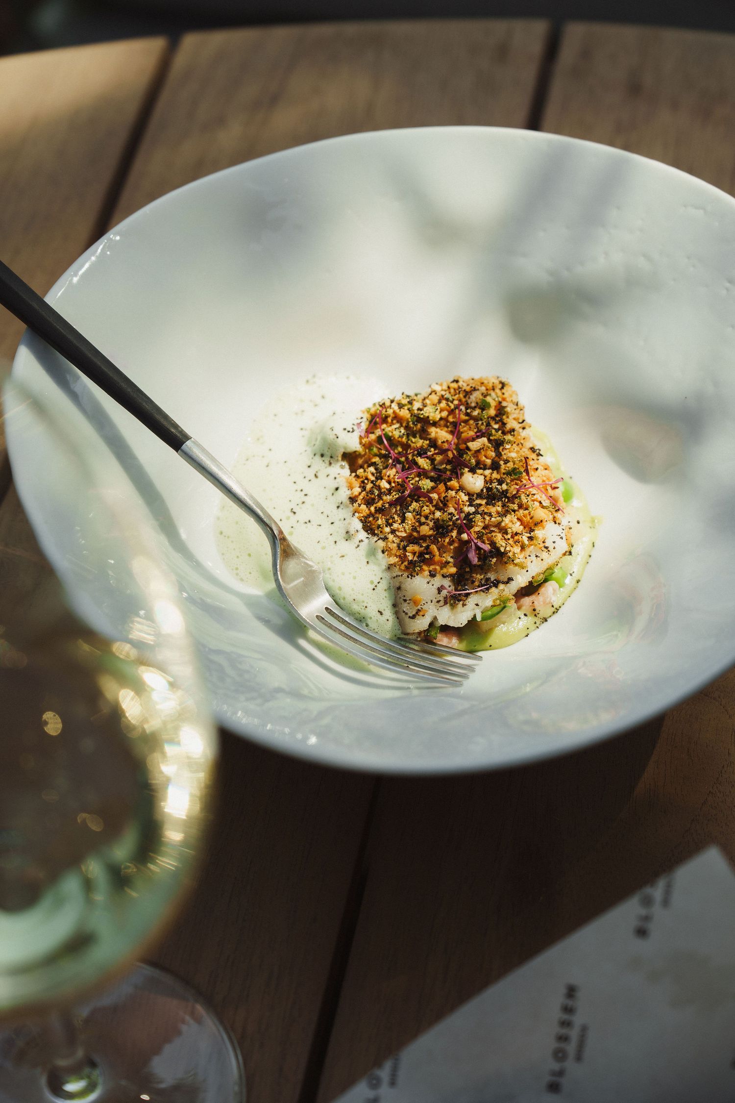 Elegant fine dining dish with herb-crusted fish on a white plate, served with a fork and white wine glass on wooden table.