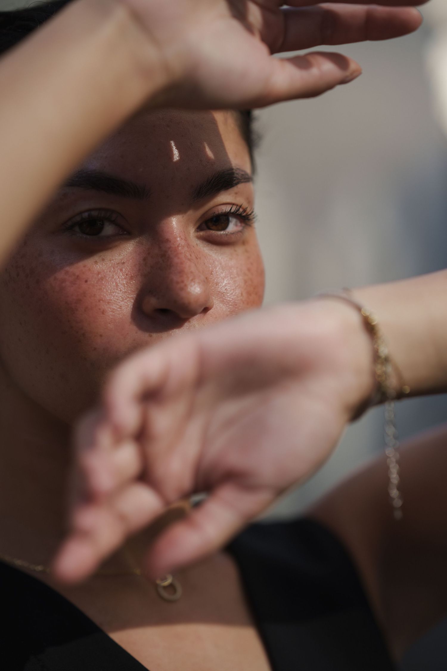 Close-up portrait of a young woman with freckles shielding her face from sunlight with both hands wearing gold bracelets.