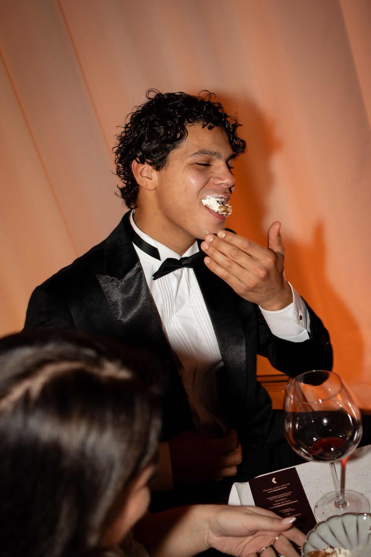 Man in tuxedo eating cake at formal dinner event with red wine glass on table.