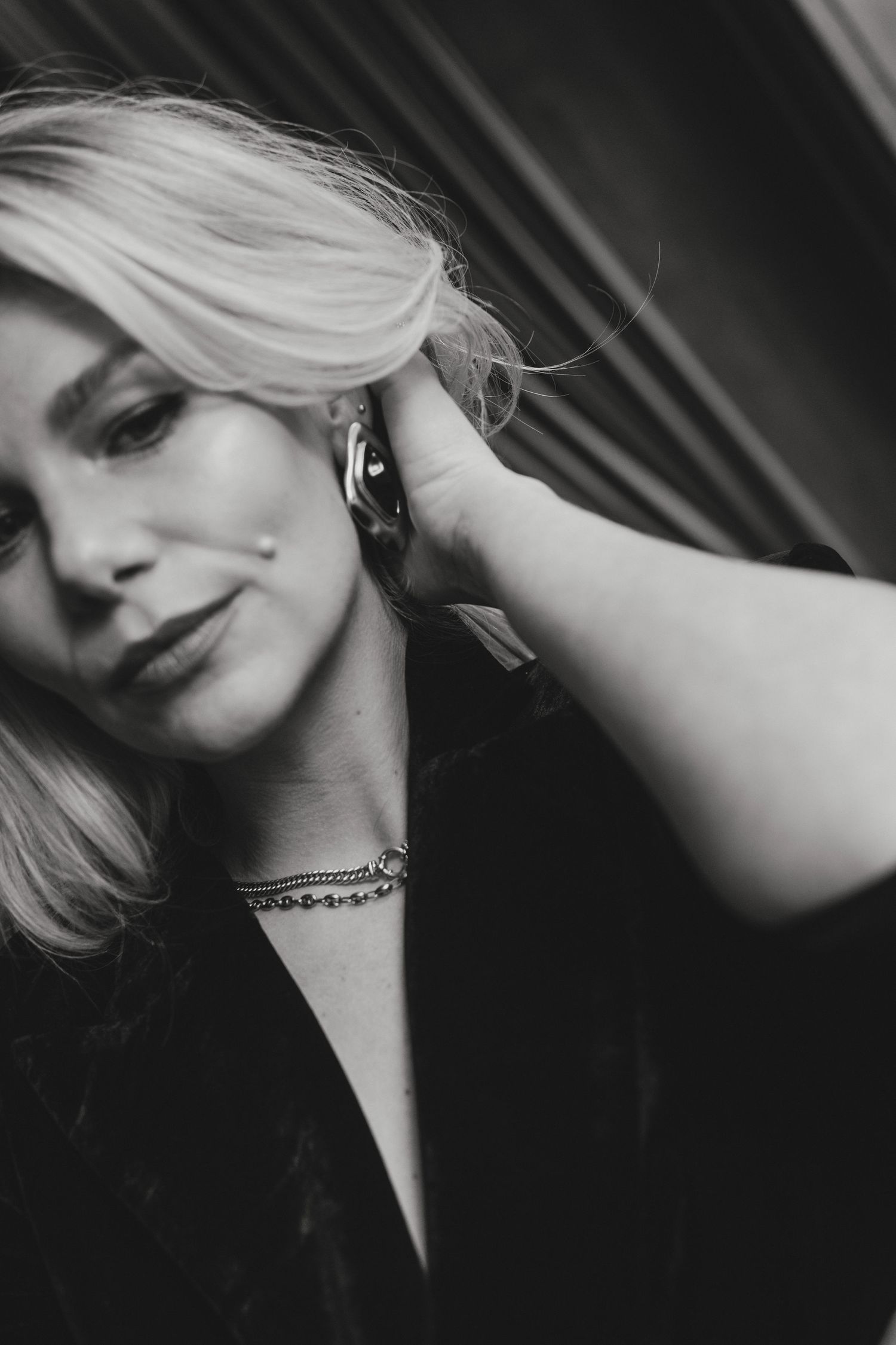 Black and white portrait of a blonde woman touching her hair, wearing hoop earrings and a chain necklace.