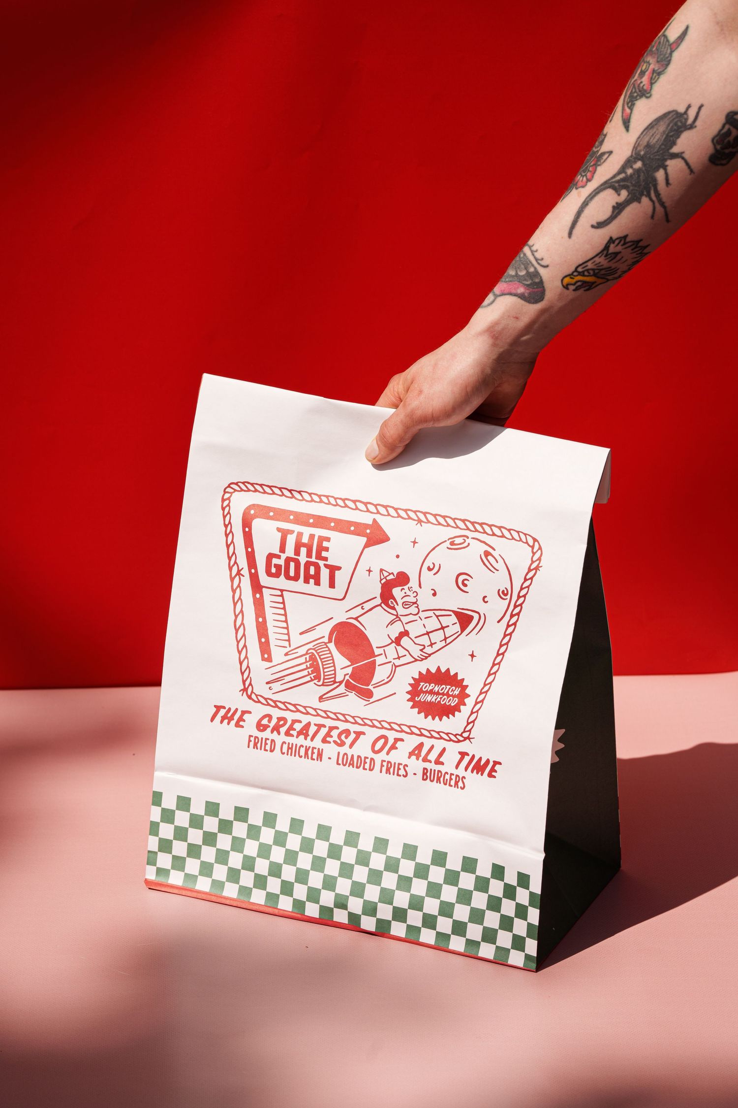 Tattooed hand holding a white paper takeout bag printed with The GOAT Greatest of All Time burger restaurant branding.