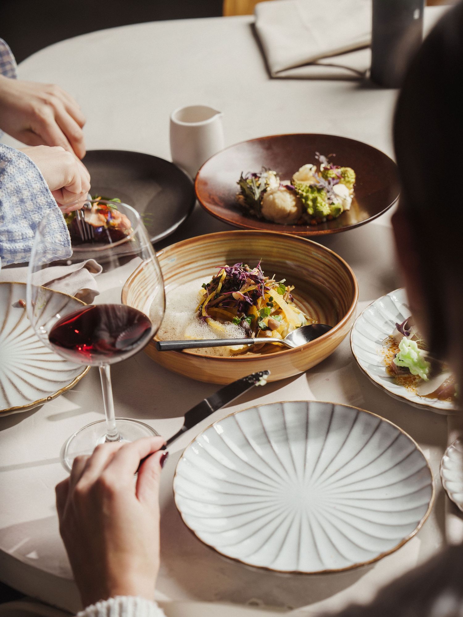 Elegant dining table with gourmet dishes, red wine glass, and ceramic plates in a sunlit upscale restaurant setting.